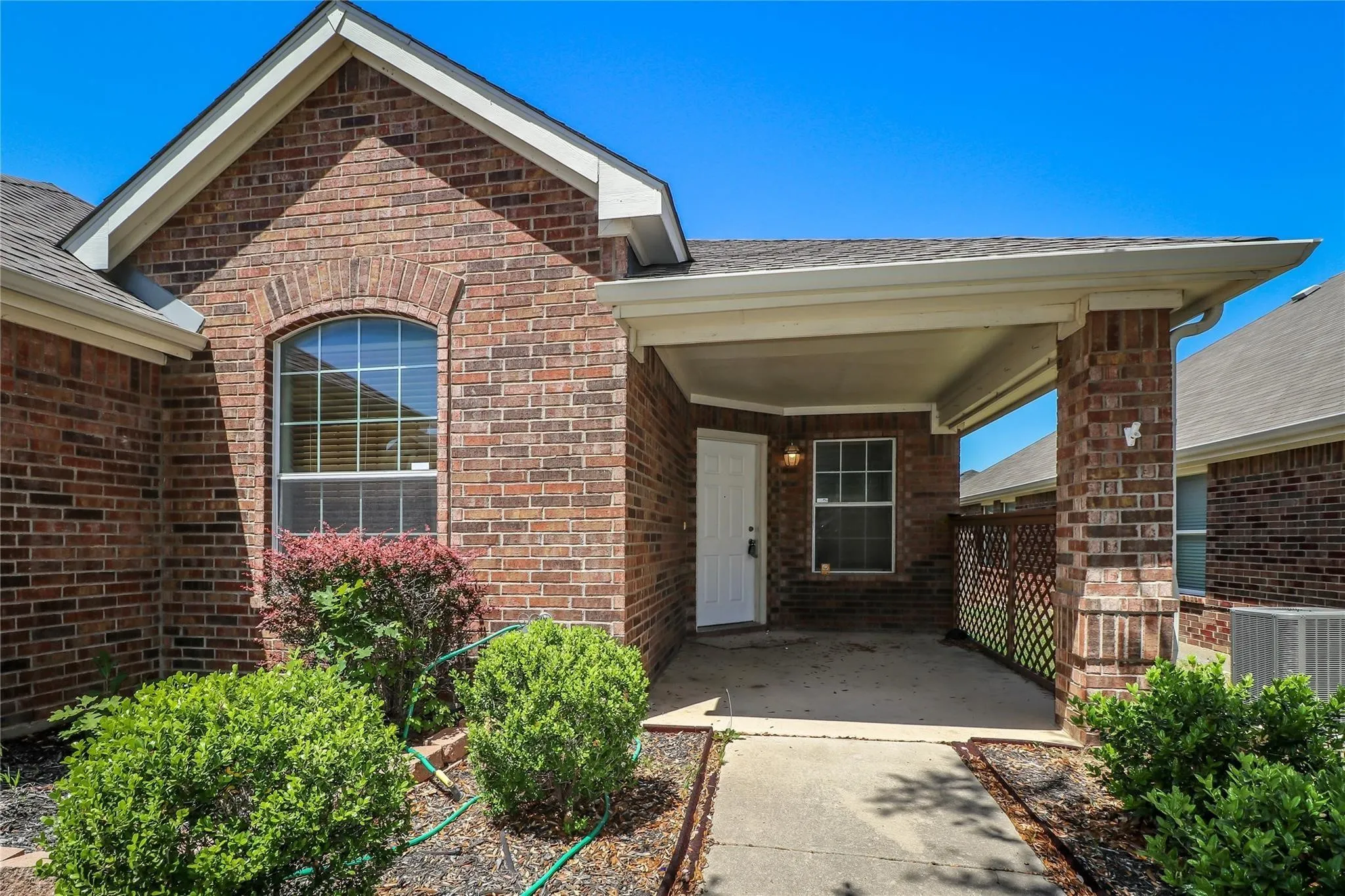 Brick exterior with an arched window, covered entryway, concrete pathway, and established plantings