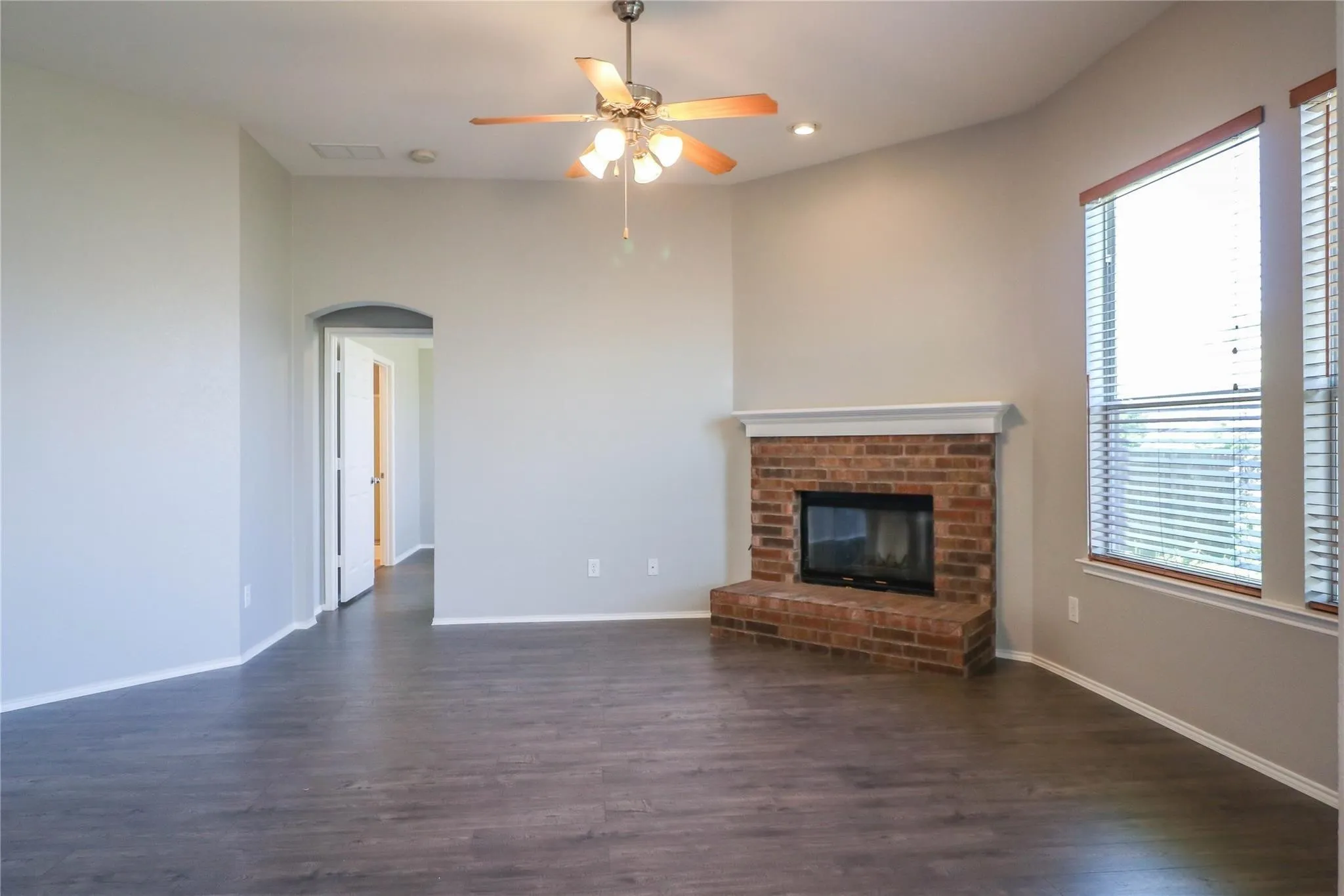 Wood-finish flooring, brick fireplace with white mantel, ceiling fan with light fixture, recessed lighting, and dual windows with blinds