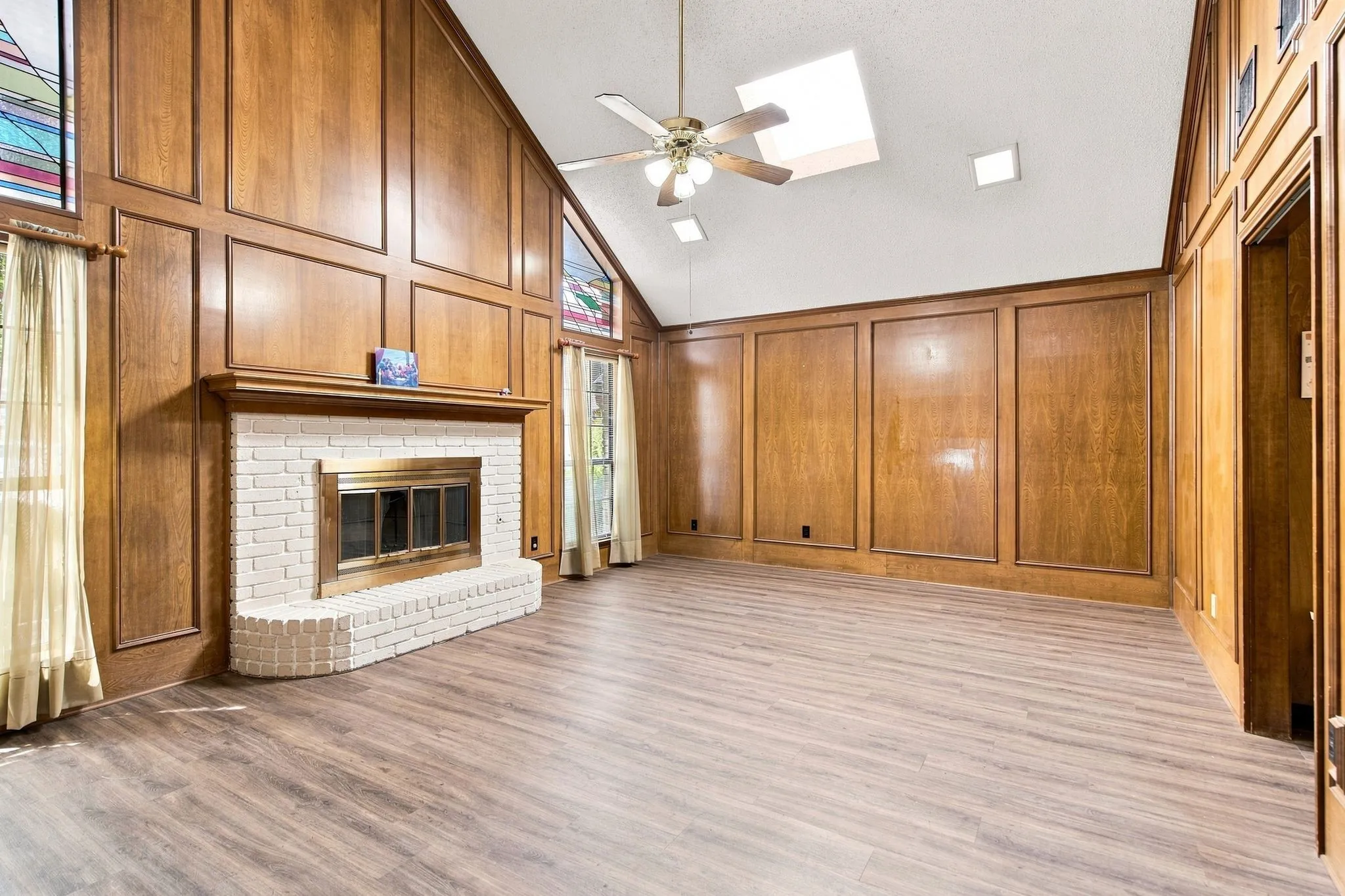 Vaulted ceiling, paneled walls, white brick fireplace with mantel, vinyl plank flooring, and ceiling fan with integrated lighting