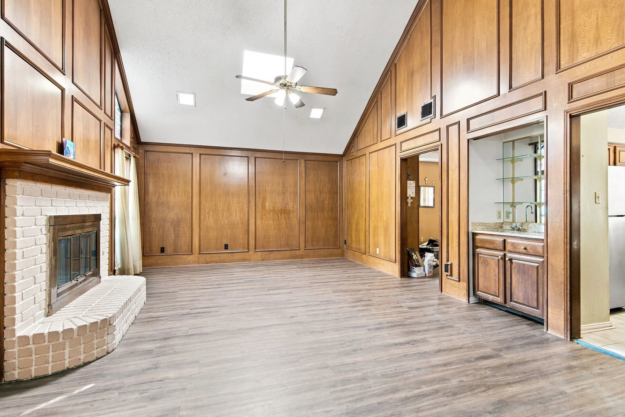 Vaulted ceiling with skylight, vinyl plank flooring, wood paneling with decorative trim, white brick fireplace, and built-in wet bar with cabinetry