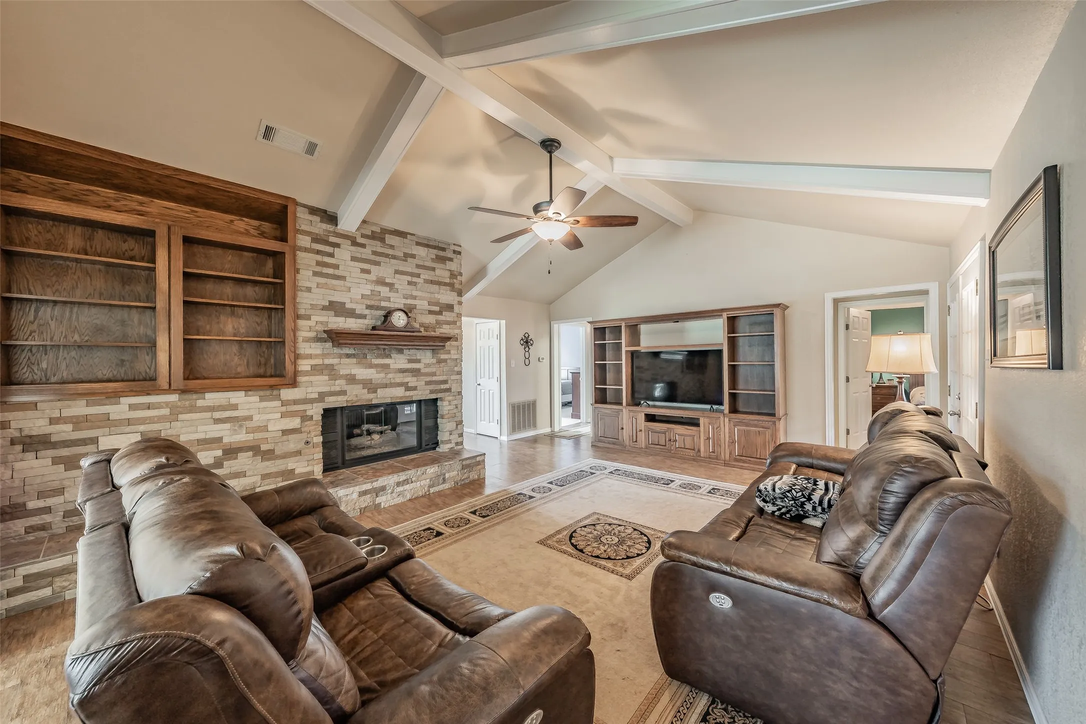 Vaulted ceiling with exposed beams, tile  flooring, stone-clad fireplace with built-in shelving, wood-finish entertainment center, and ceiling fan