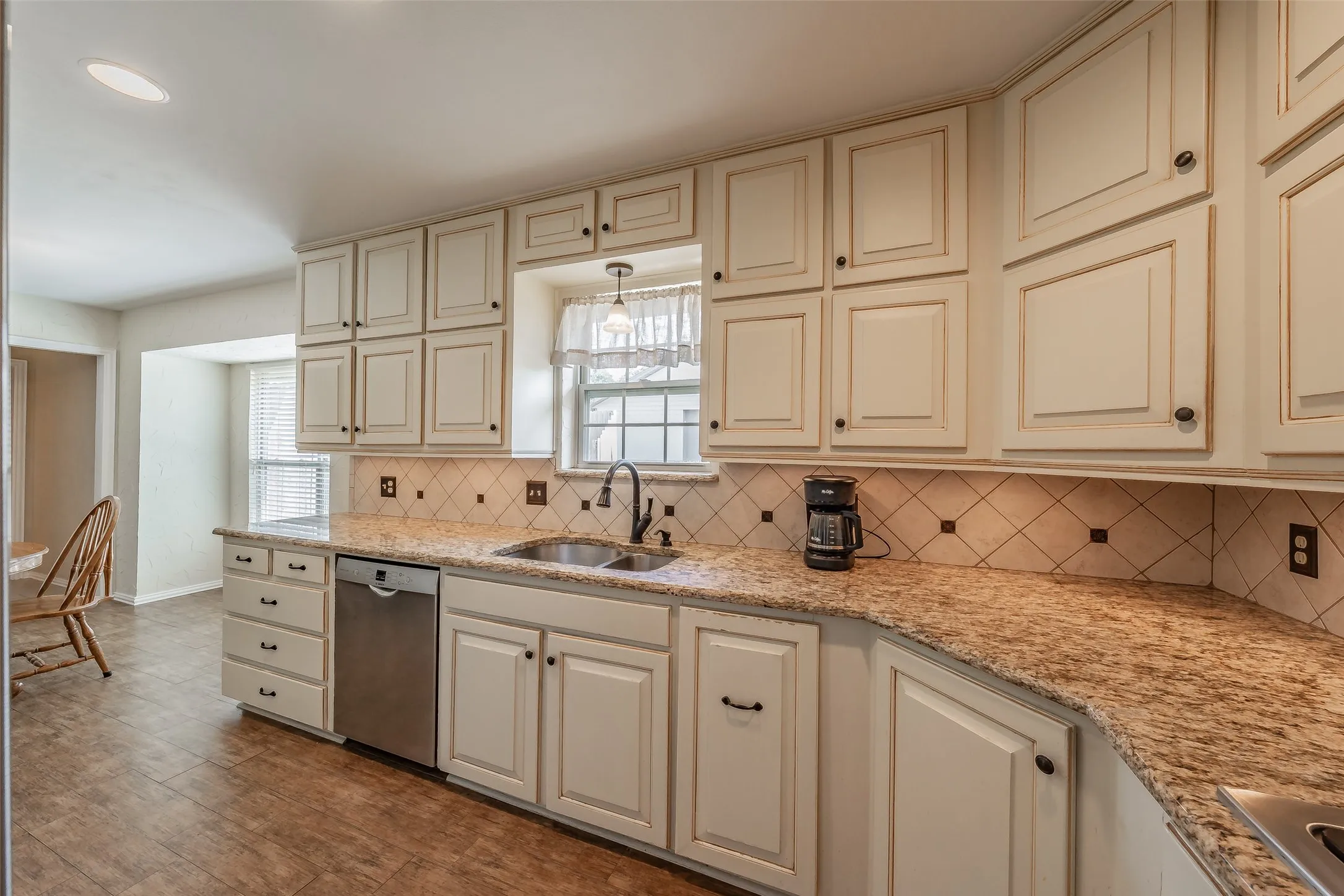 Cream-finish cabinetry with dark hardware, granite-style countertops, stainless steel dishwasher, tile backsplash, and tile flooring