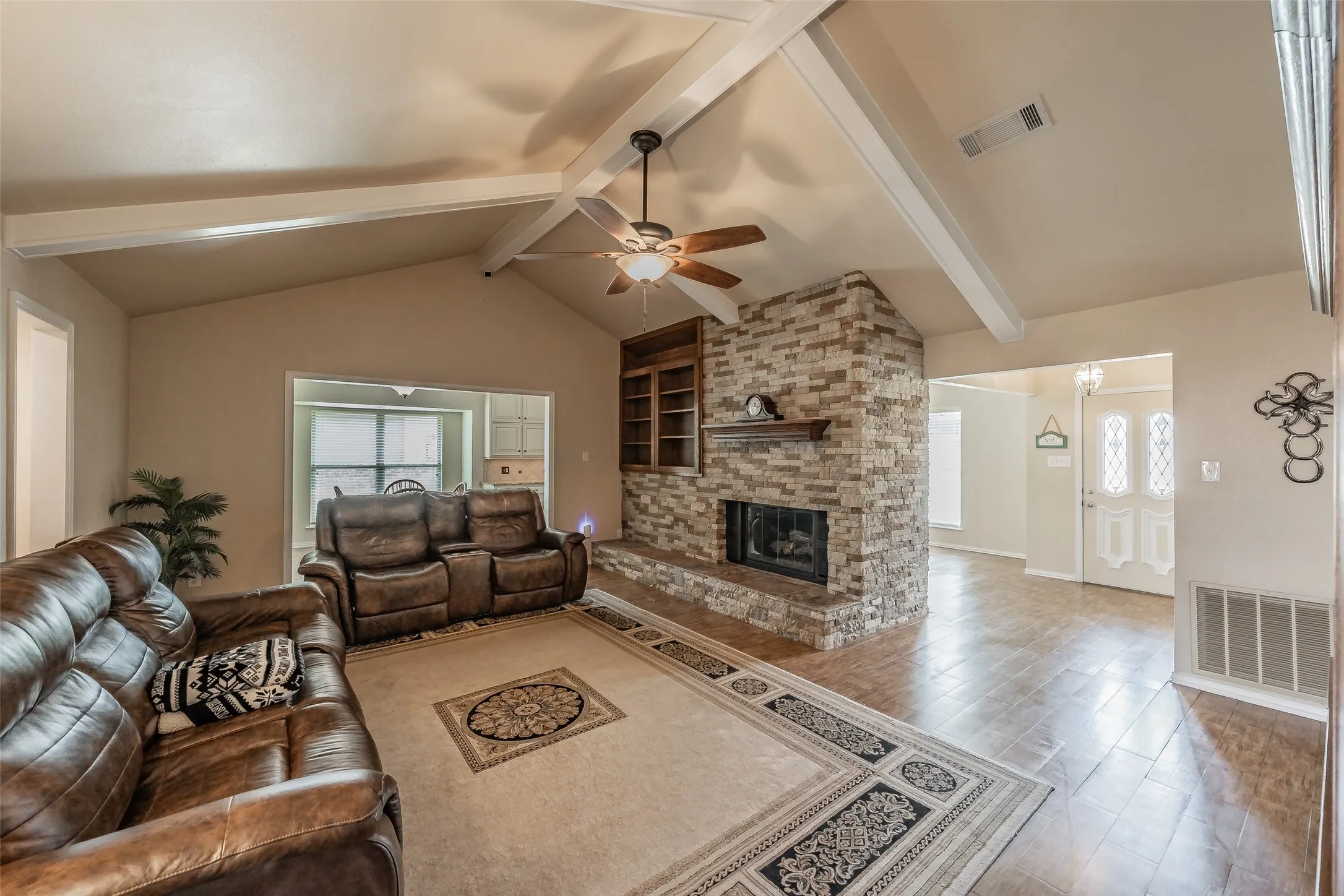 Vaulted ceiling with exposed beams, stone-faced fireplace with hearth, built-in shelving, wood-finish flooring, and a decorative front entry door