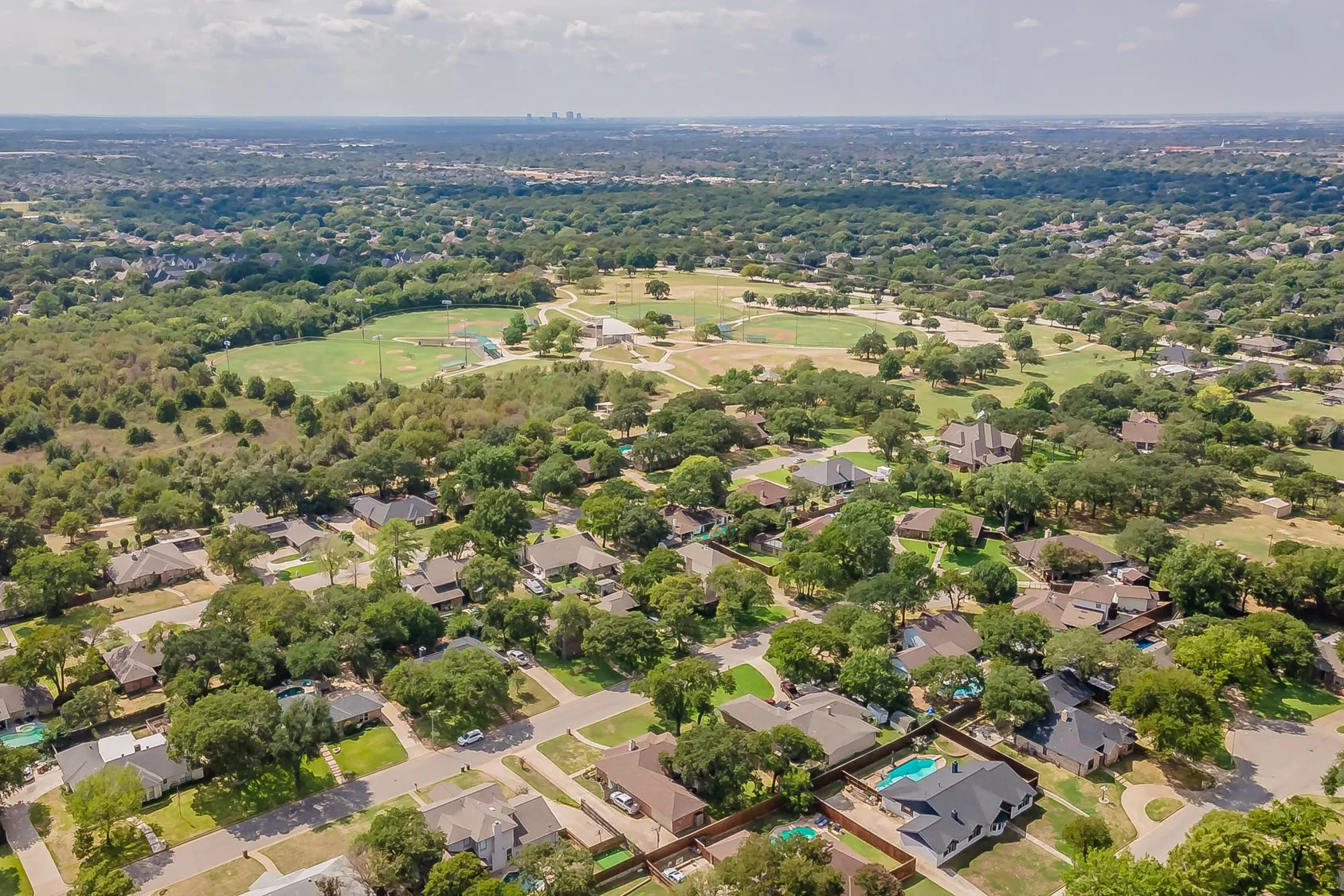 Aerial perspective of a residential area featuring mature tree coverage, a large green space, and multiple properties with private pools