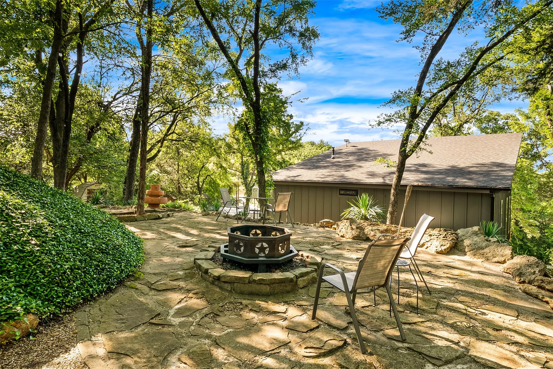 Stone patio with a built-in fire pit, surrounded by lush landscaping and mature trees