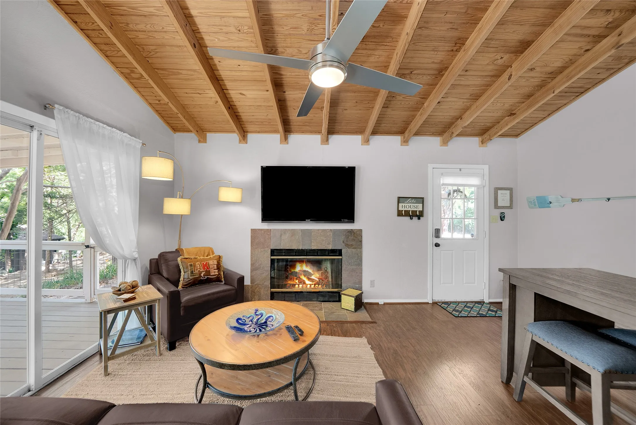 Wood-finish flooring with a wood-beamed ceiling, stone fireplace, and a ceiling fan with integrated lighting