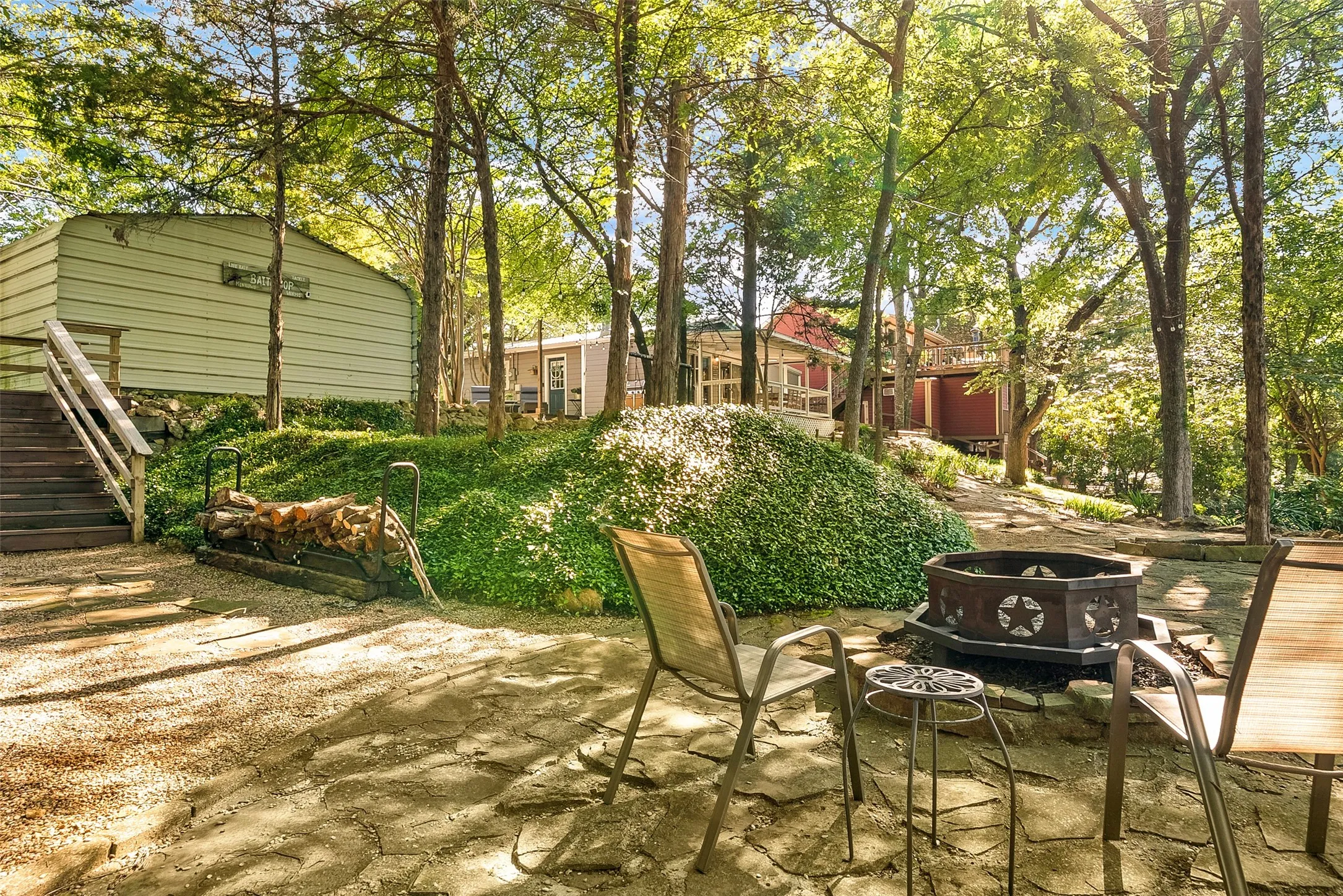 Stone patio with a decorative fire pit, surrounded by mature trees and lush ground cover