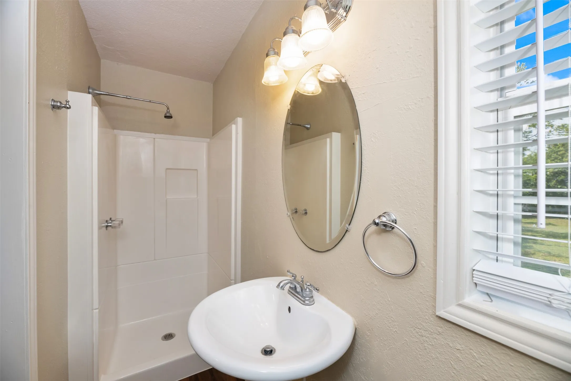 Pedestal sink with chrome faucet, oval mirror, wall-mounted lighting fixture, and a white framed window with horizontal blinds