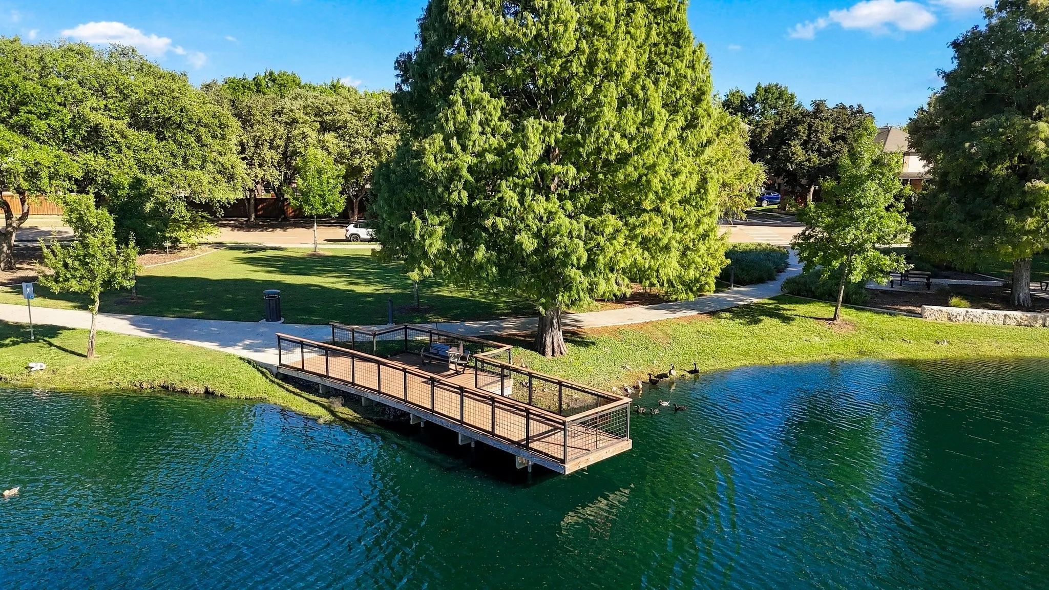 Waterfront property featuring a wooden deck with dark railings, a paved pathway, and mature trees