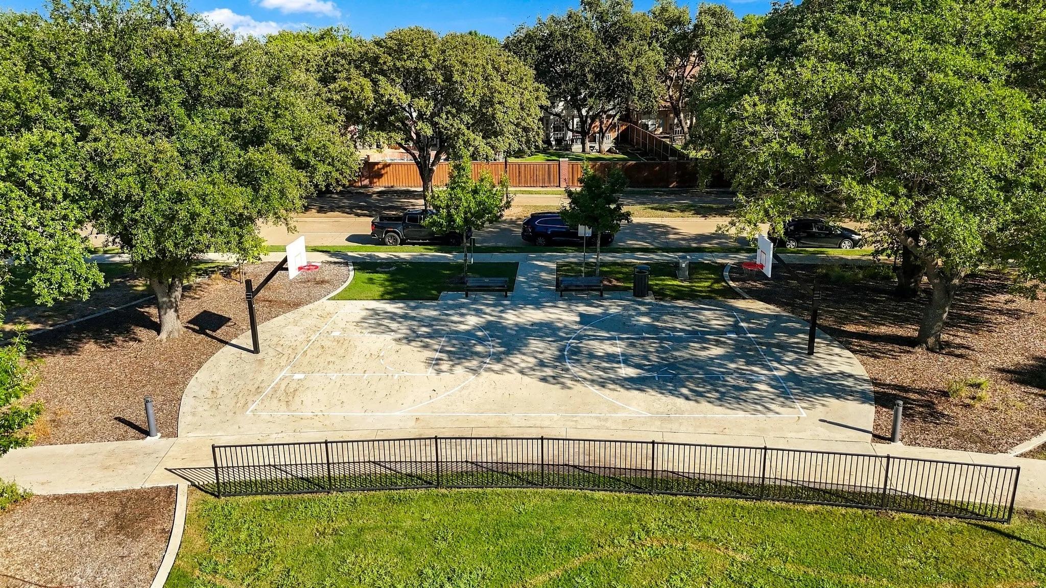 Outdoor basketball court with two hoops, surrounded by mature trees and a manicured lawn