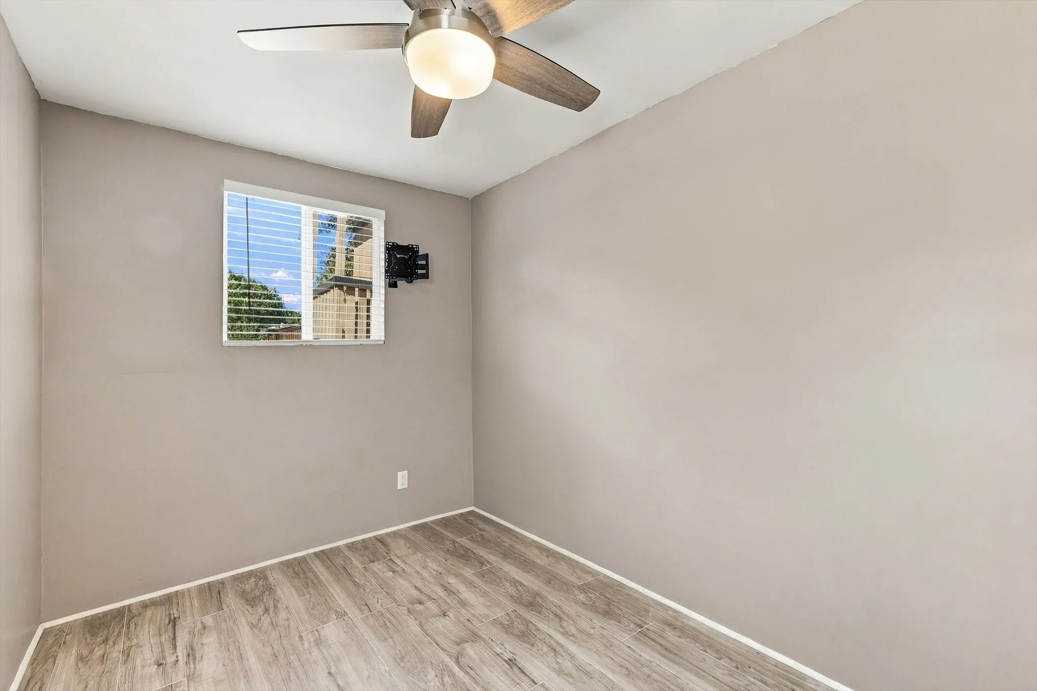 Wood-finish flooring, neutral wall paint, window with blinds, ceiling fan with integrated lighting, and a wall-mounted bracket