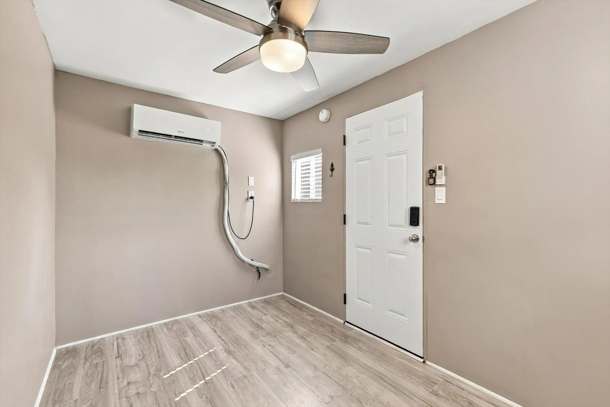 Wood-finish flooring, ceiling fan with integrated lighting, wall-mounted air conditioning unit, white paneled door, and horizontal window blinds