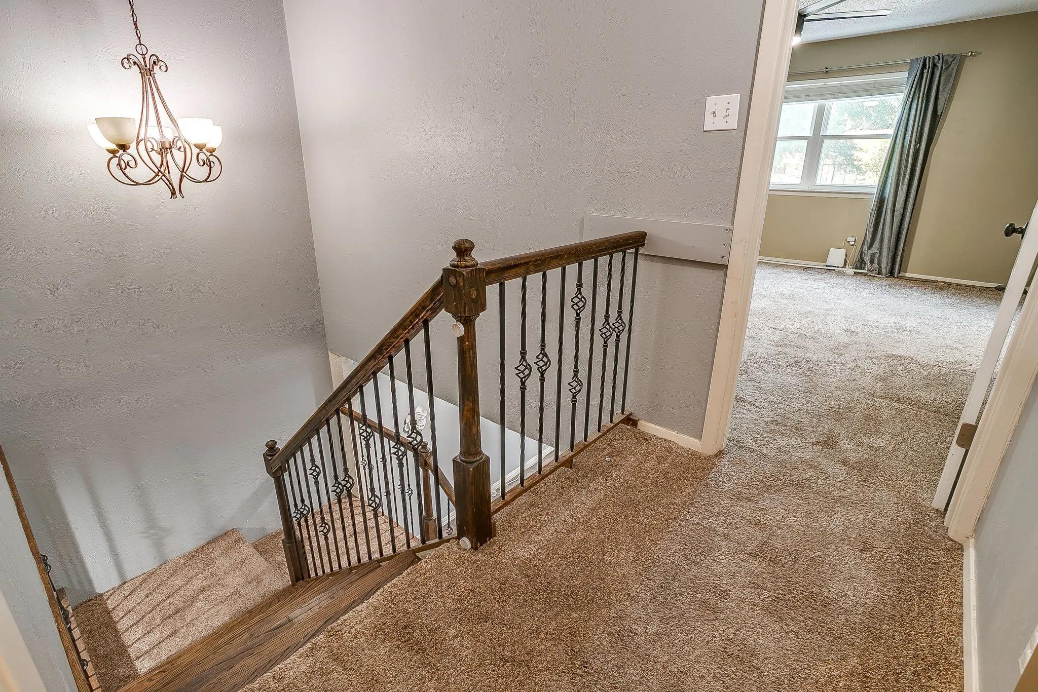 Carpeted staircase with dark wood-finish handrail and ornate iron balusters, illuminated by a decorative chandelier