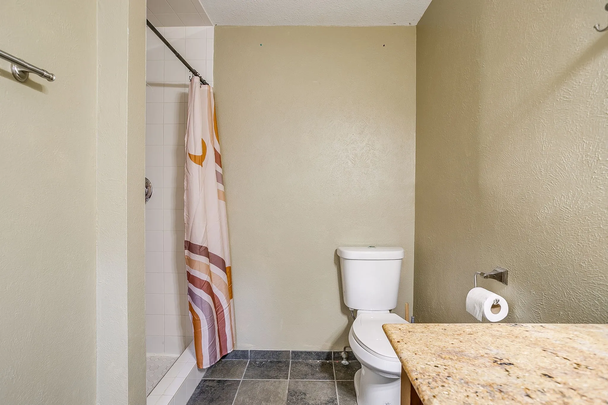 Shower with white tile surround, toilet, and vanity with stone-finish countertop