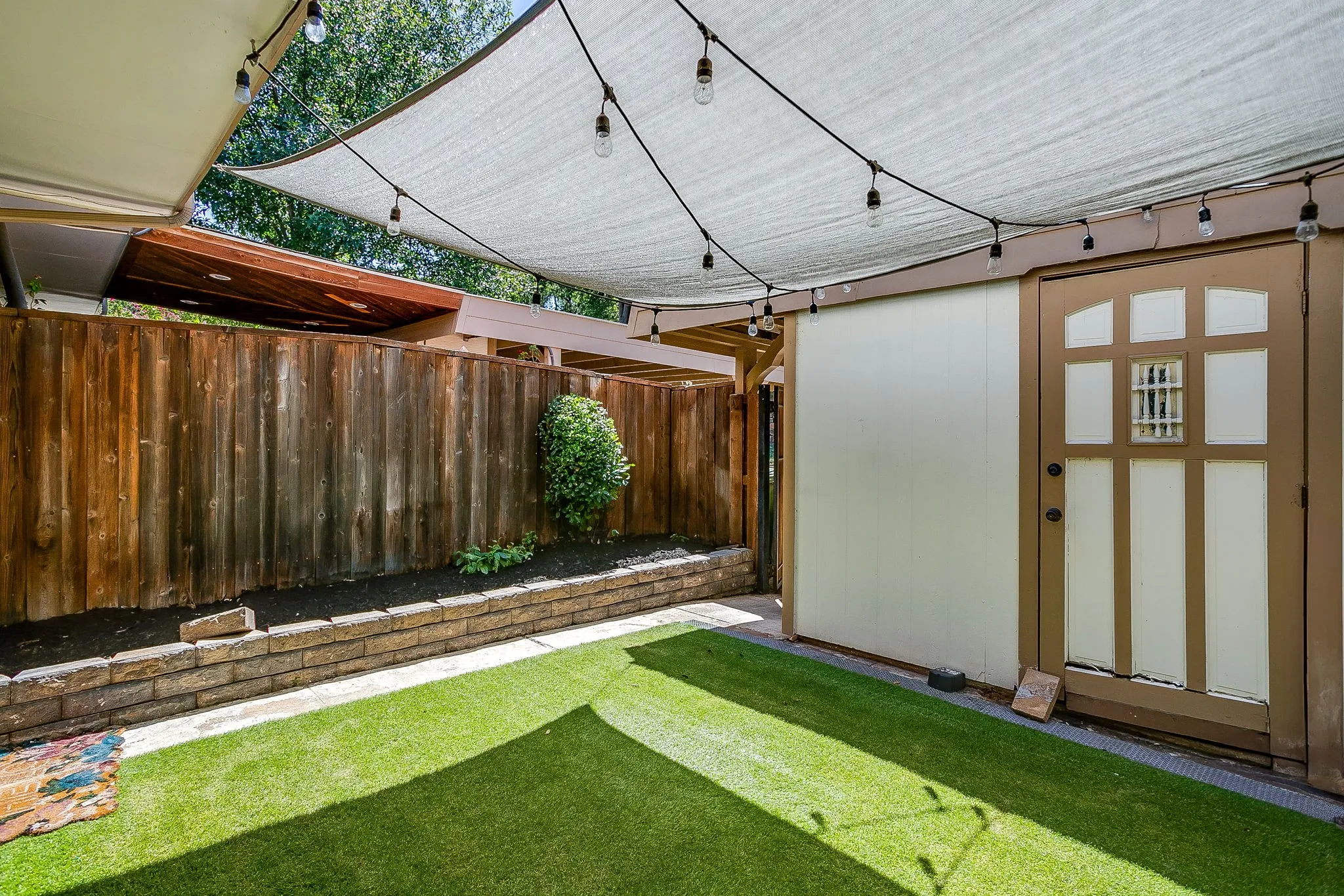 Fenced outdoor area featuring artificial turf, a stone retaining wall, and a shade sail with string lighting