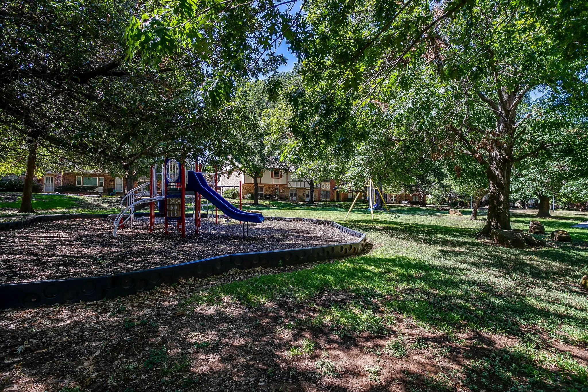 Recreational area featuring a playground structure with slides, swing set, and mature trees providing shade