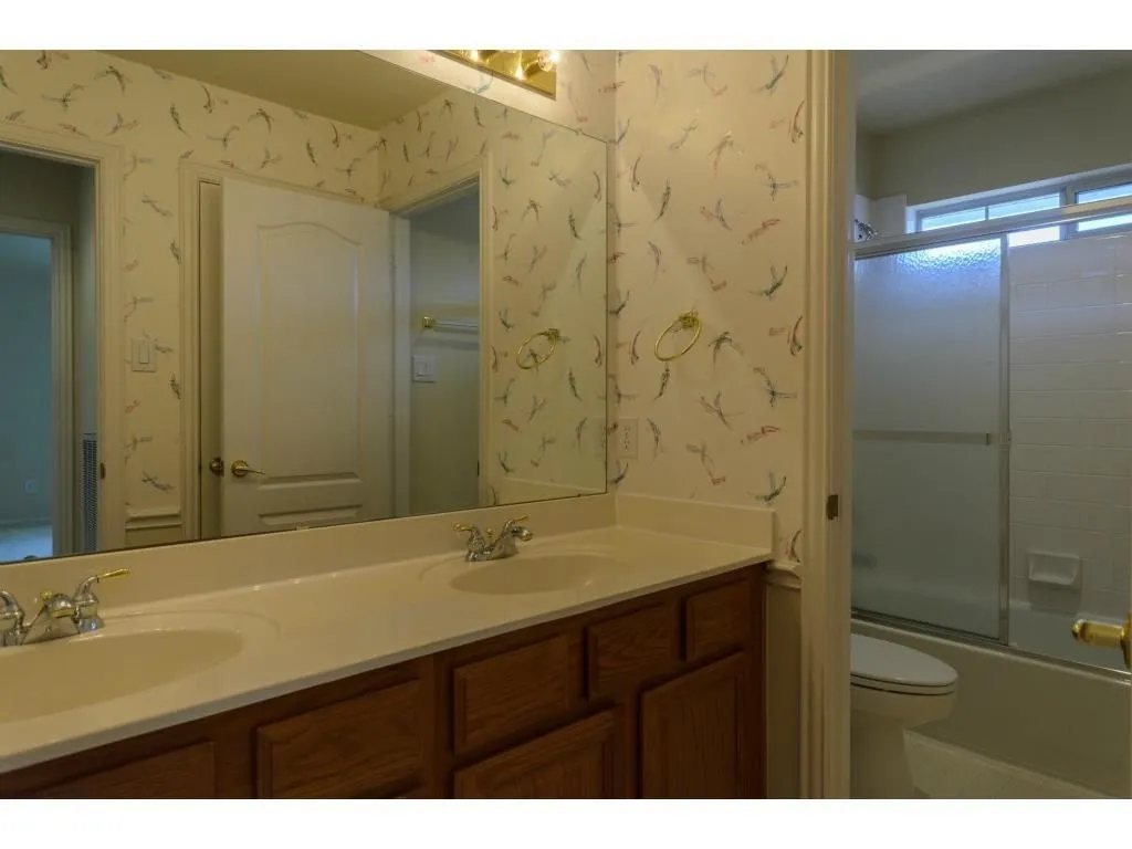 Dual basin vanity with chrome and gold-tone fixtures, wood-finish cabinetry, and full-width mirror
