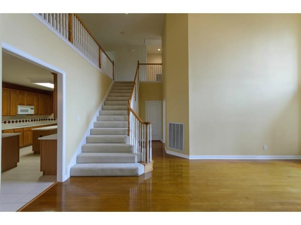 Hardwood flooring, carpeted staircase with wood banister and white balusters, overhead lighting, kitchen with wood cabinetry and tile backsplash, double-height wall