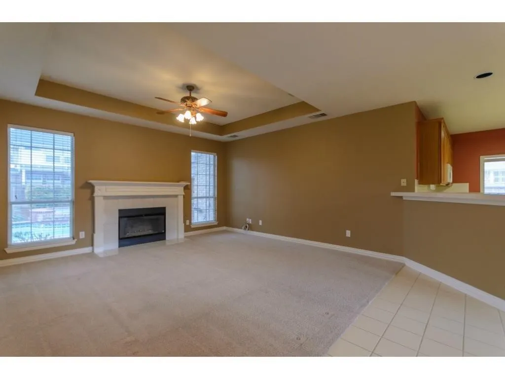Recessed ceiling with a central fan light fixture, gas fireplace with a white mantel, light-colored carpeting, two large windows, and a tiled floor transition
