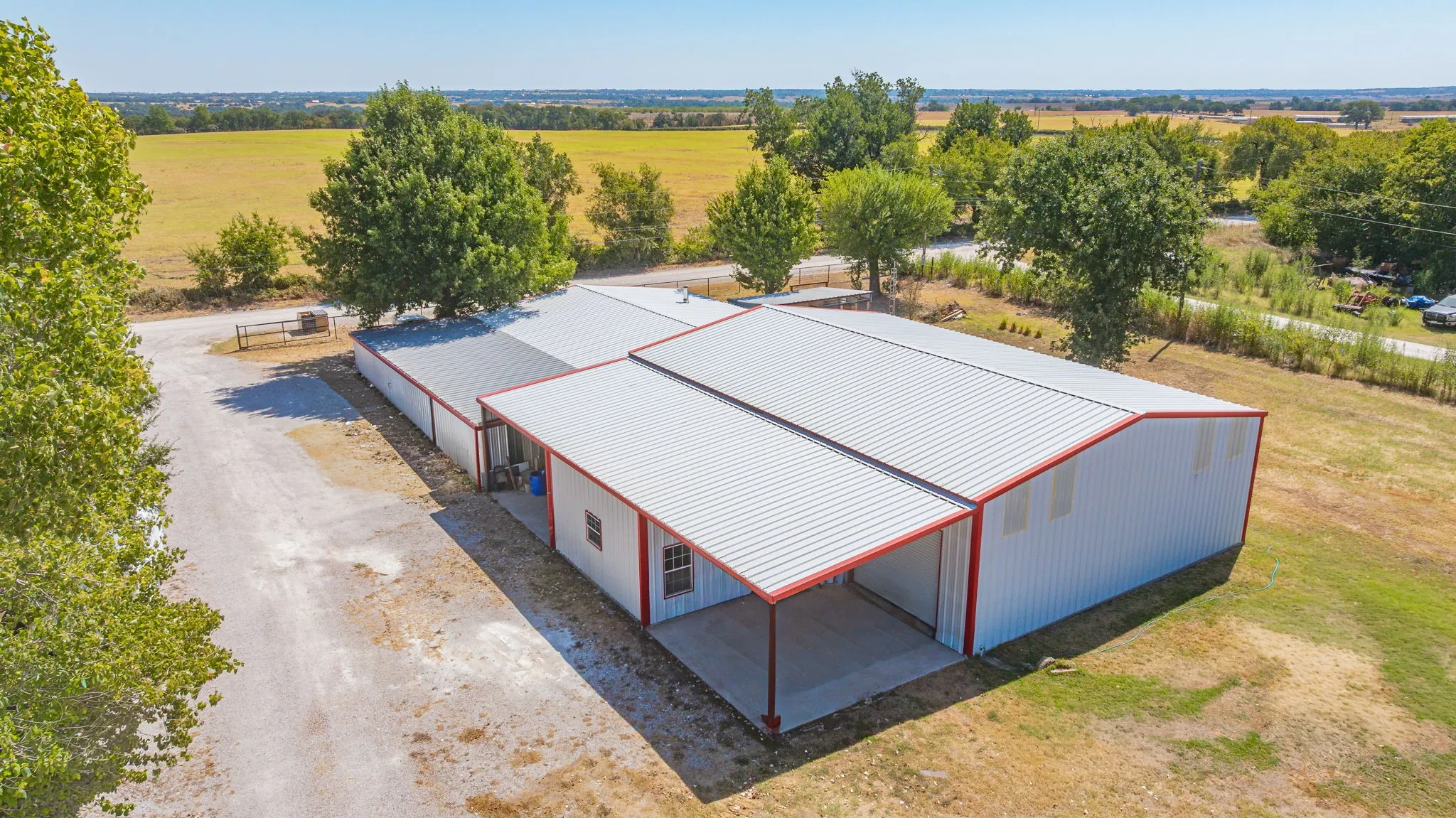 Expansive metal structure featuring a white exterior with red trim, a covered concrete patio, and a large gravel driveway