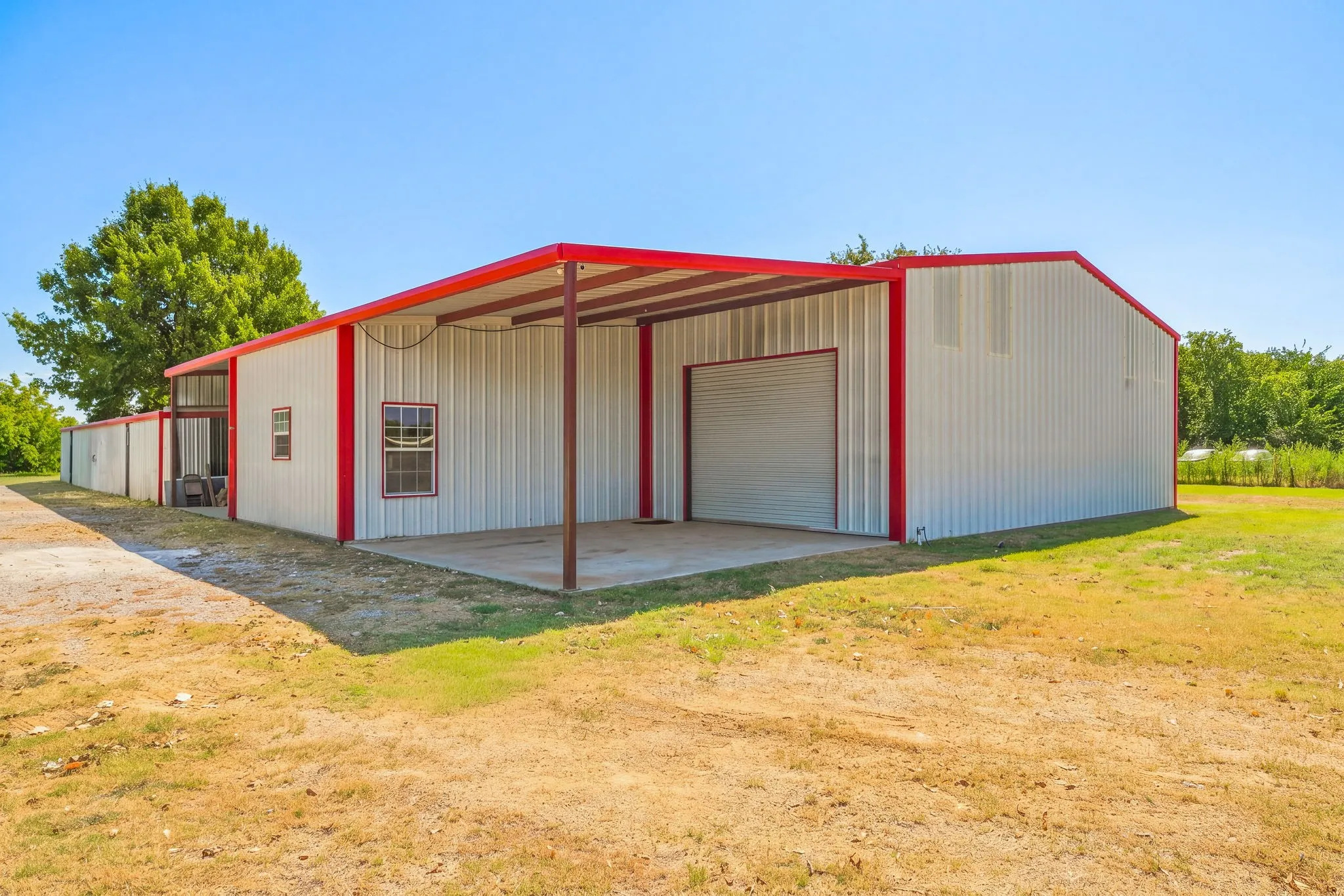 Metal building featuring a red trim, multiple windows, a covered concrete pad, and a large roll-up door