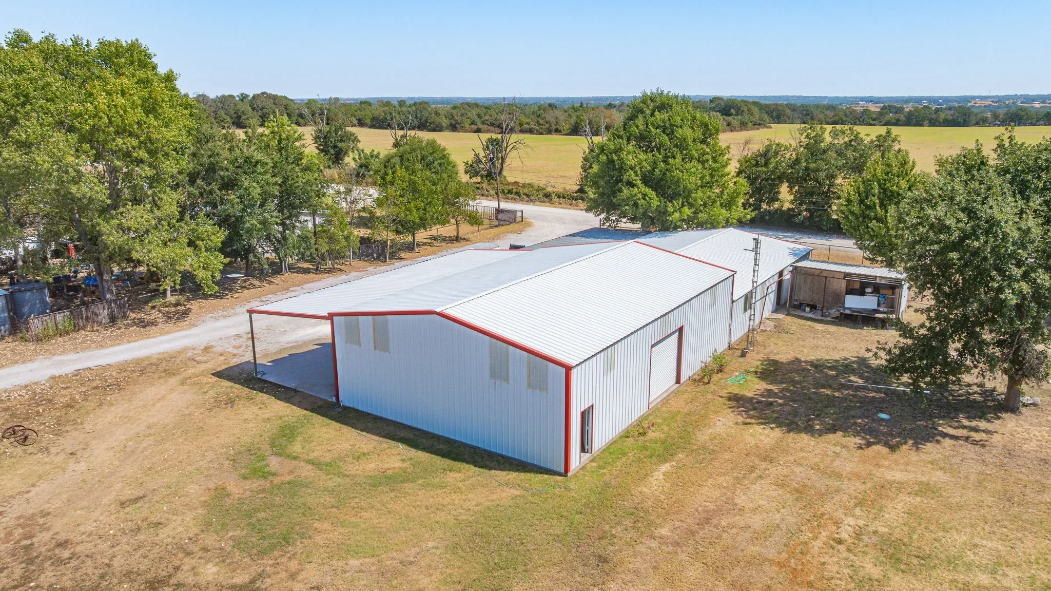 White metal building with red trim, attached covered parking, gravel drive, and adjacent outbuilding