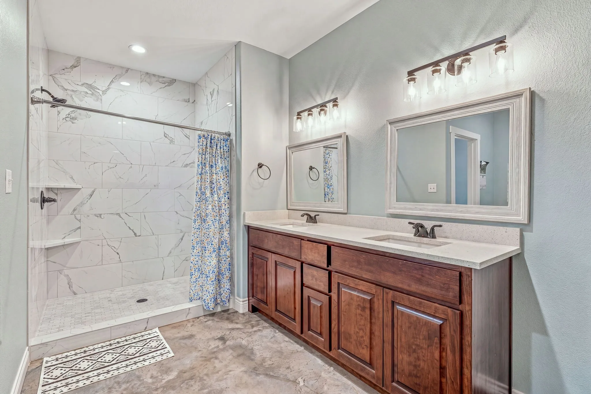 Walk-in shower with tiled walls and floor, dual vanity with wood cabinetry, light-colored countertops, and two framed mirrors