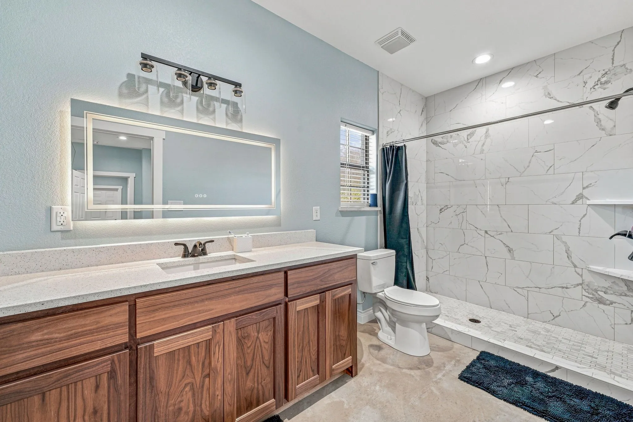 Spacious bathroom featuring an elongated wood-finish vanity with an integrated sink and light-toned countertop, a large illuminated mirror, and a walk-in shower with white tiled walls and a built-in niche