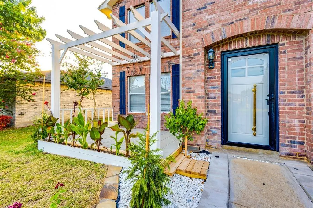 Brick exterior with arched entryway, a white pergola, a raised planting bed, a front door with decorative glass, and blue window shutters