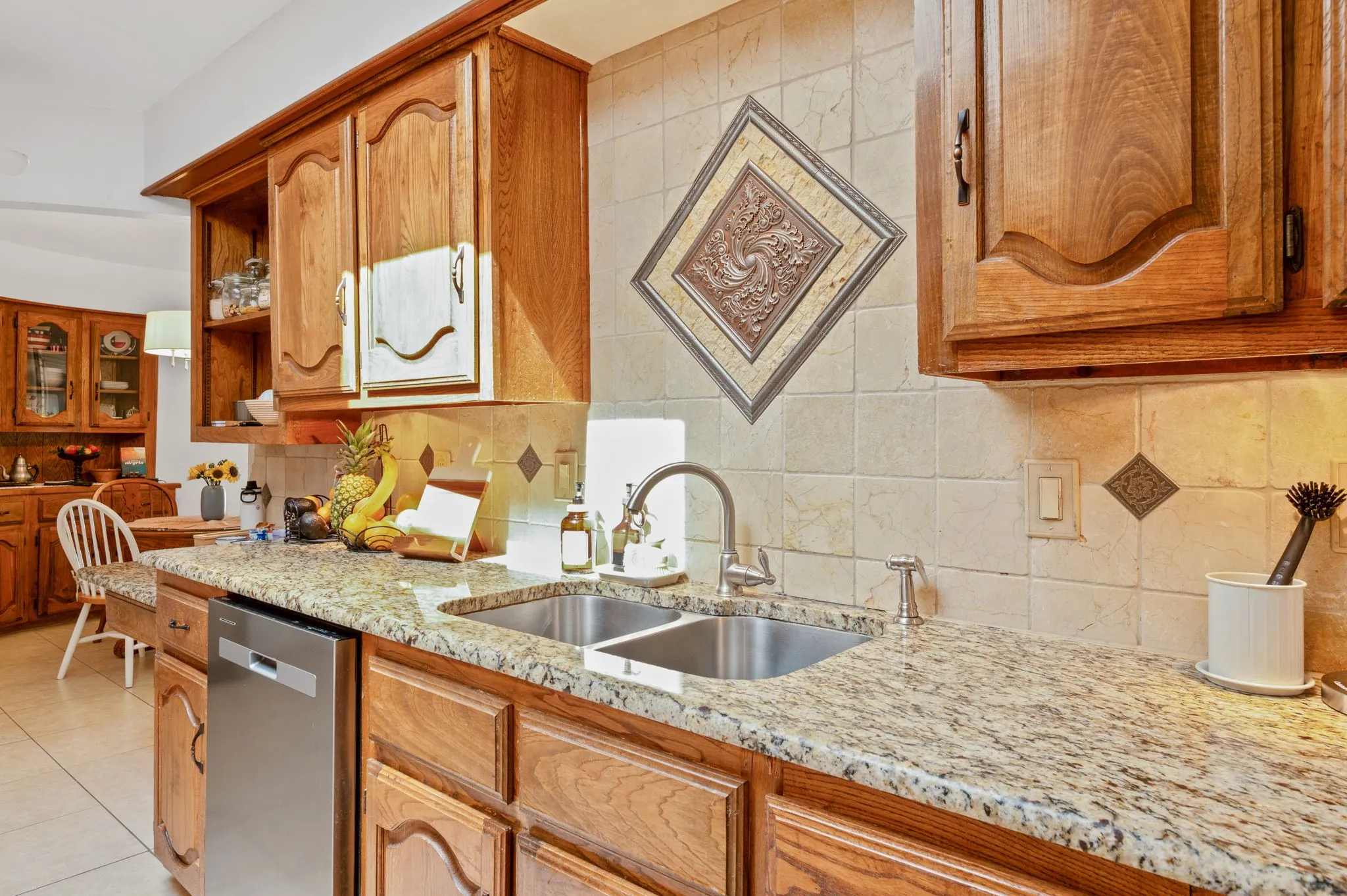 Granite-style countertops, dual stainless steel sink, tile backsplash with decorative accents, wood cabinetry, and stainless steel dishwasher
