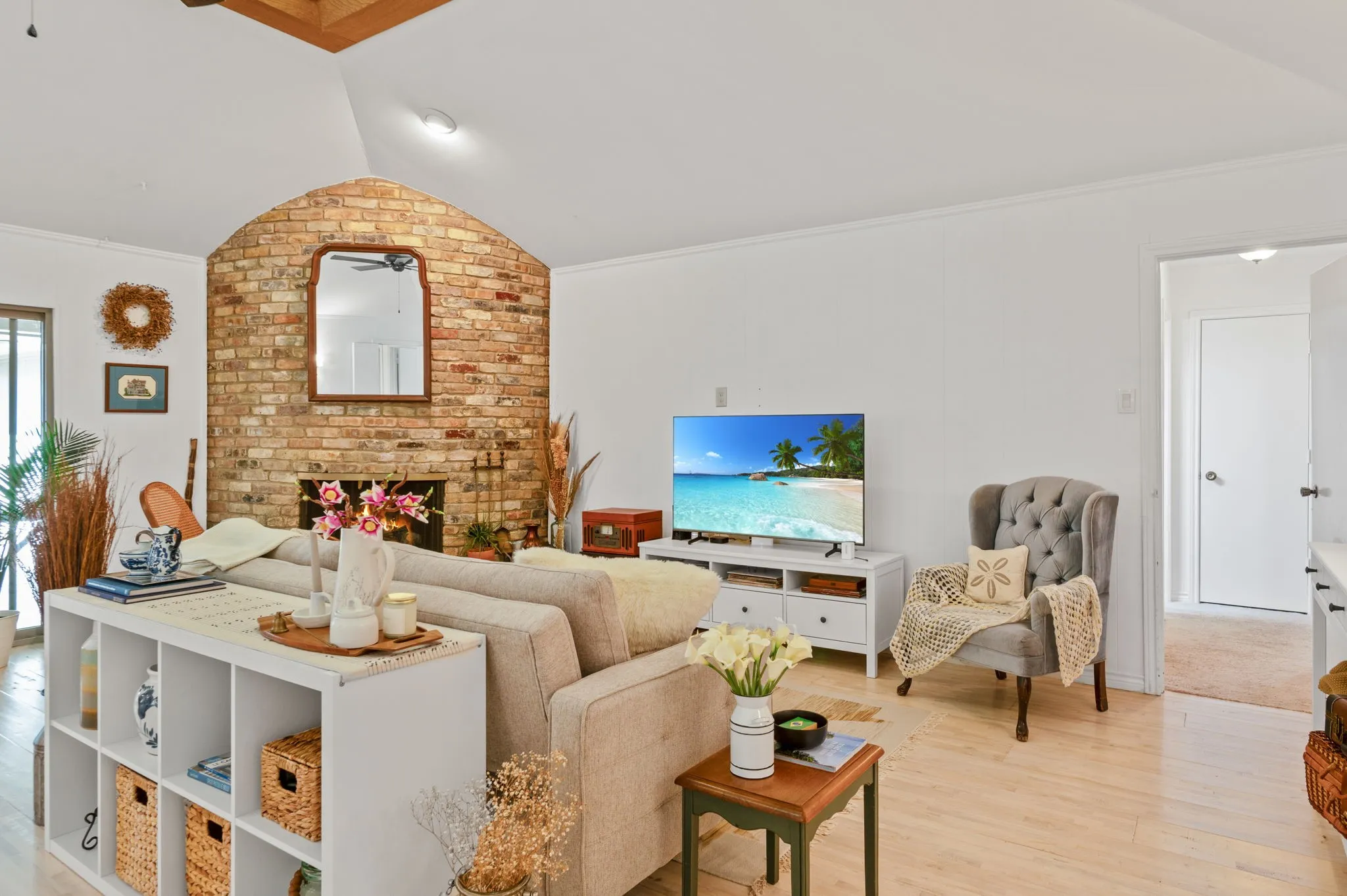 Vaulted ceiling with wood-finish beam, brick fireplace with arched detailing, light wood-finish flooring, and white painted walls