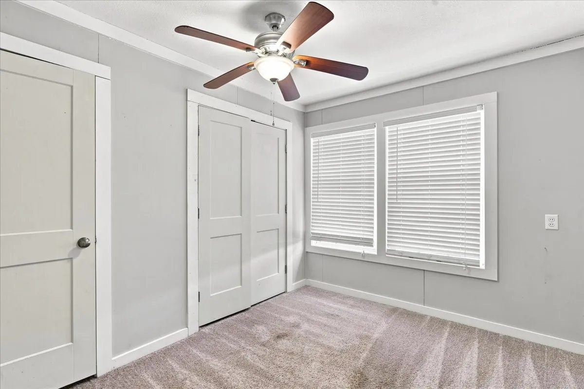 Light gray walls with white trim, a ceiling fan with wood-finish blades, dual windows with blinds, and neutral-toned carpet