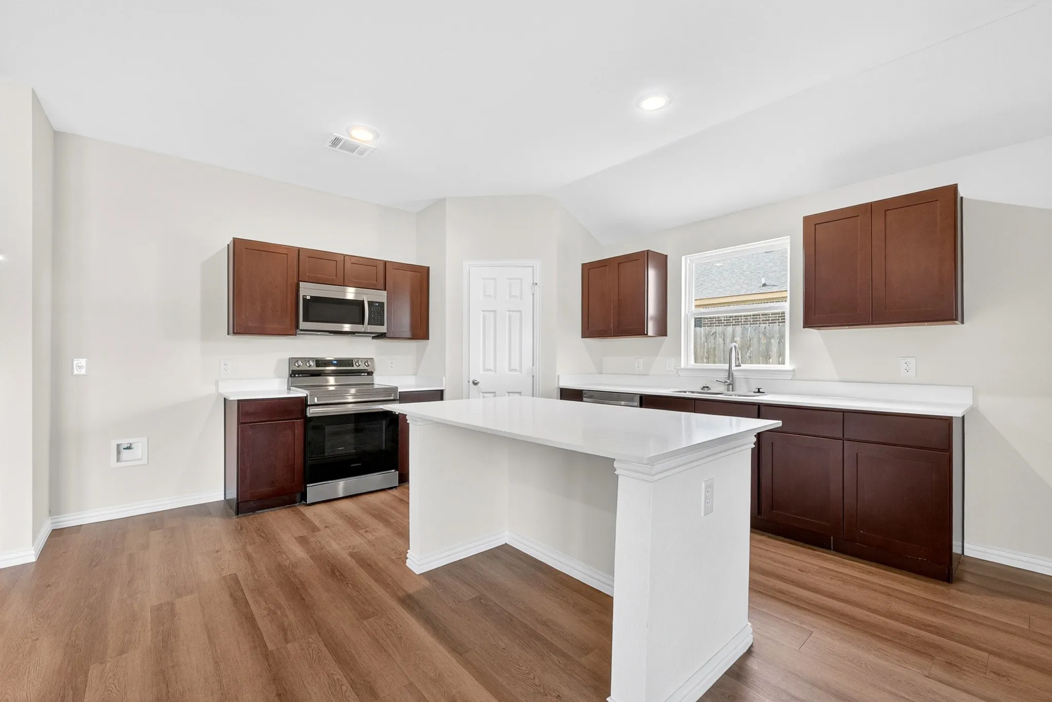 Kitchen featuring wood-finish flooring, a central island with white countertop, dark wood-finish cabinetry, stainless steel appliances, and a window above the sink