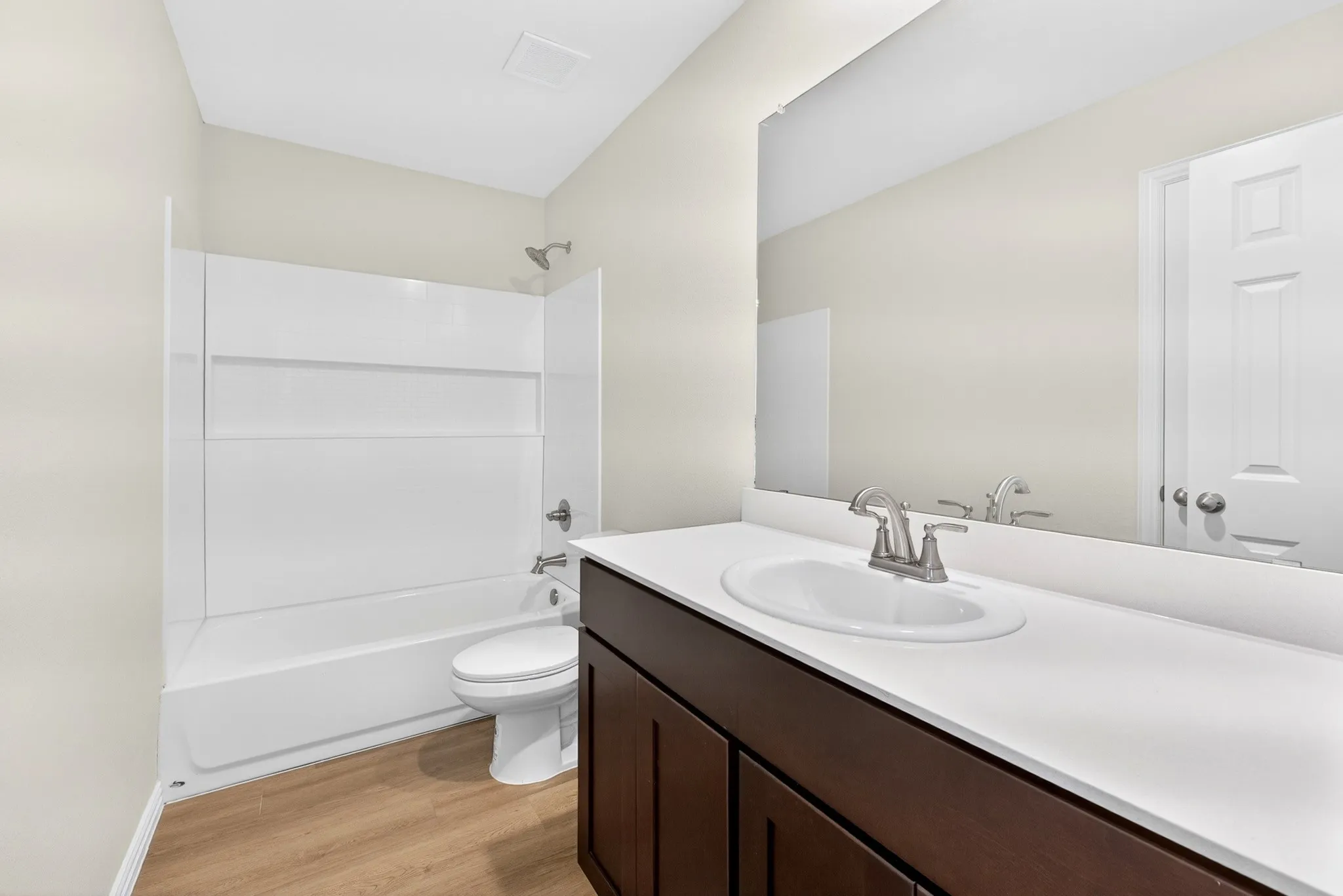 Wood-finish flooring, dark wood-finish vanity, white countertop with integrated sink, brushed nickel faucet, and a combination tub/shower with white surround