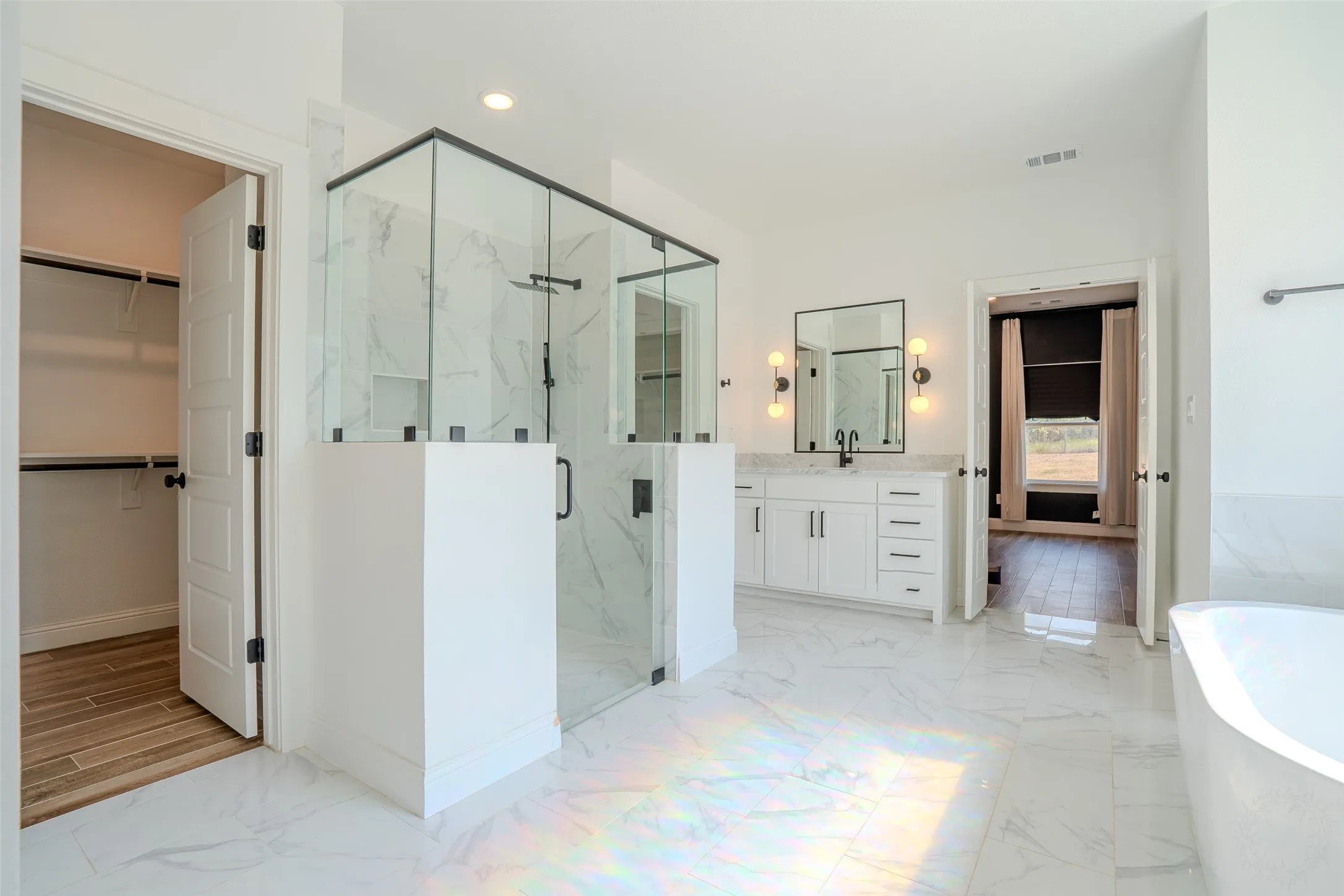 Spacious bathroom featuring a glass-enclosed shower, a double vanity with a rectangular mirror and sconce lighting, and a built-in soaker bathtub. Window directly above tub provides an abundance of natural light which makes this room a joyful experience.