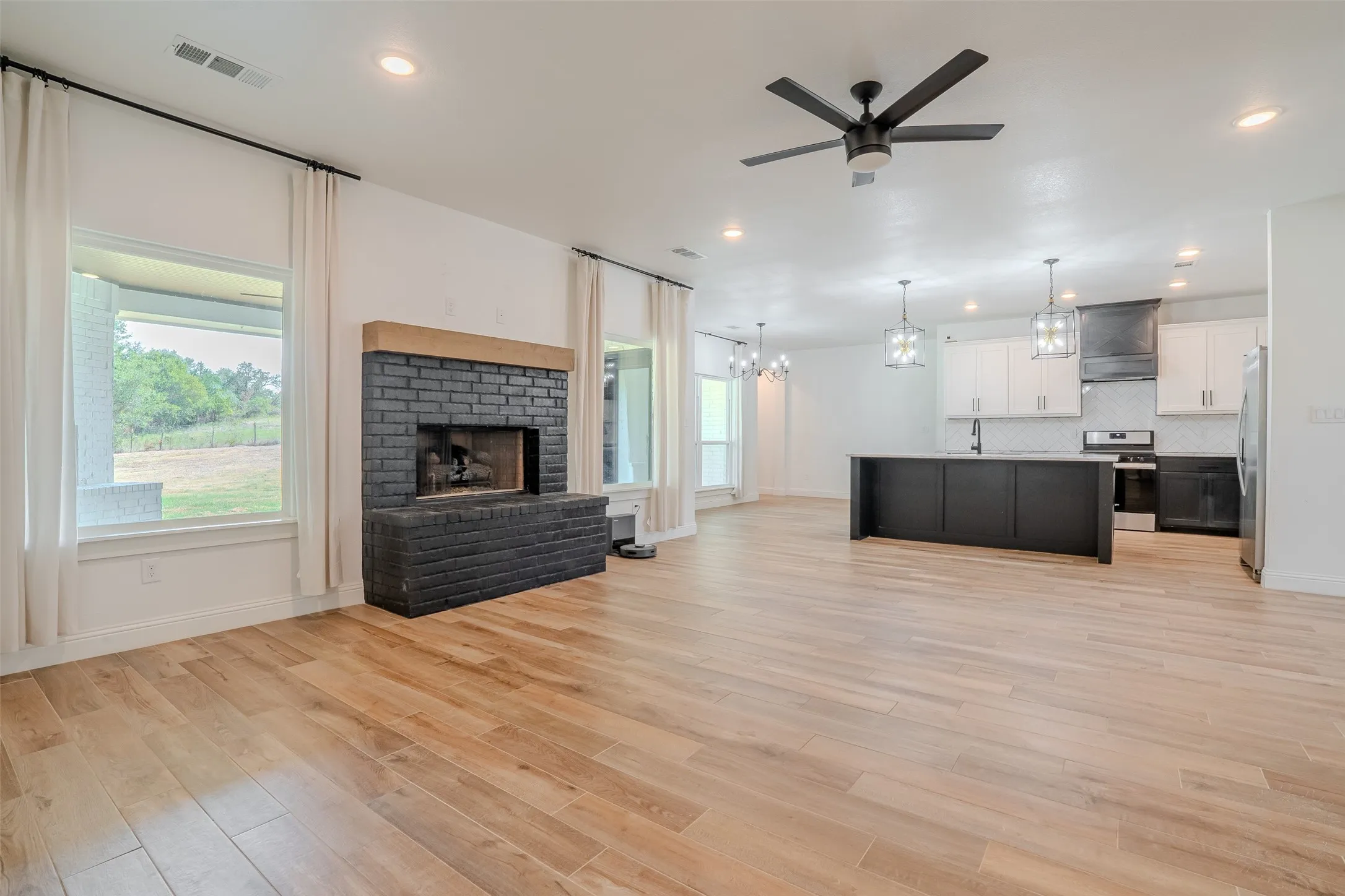 Expansive open-concept space featuring wood-look tileflooring, a black brick fireplace with a wood mantle, a modern ceiling fan, and recessed lighting