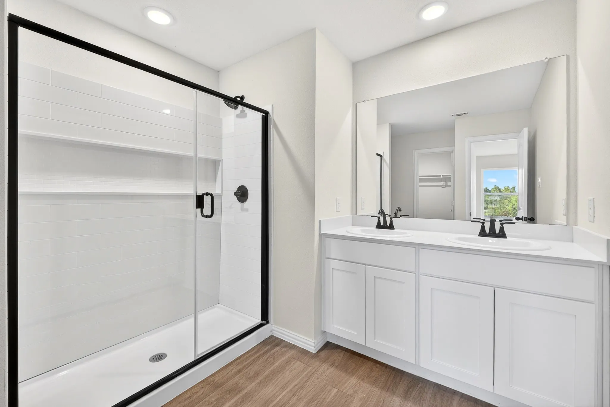 Dual vanity with white cabinetry and integrated sinks, matte black fixtures, frameless wall mirror, walk-in shower with clear glass enclosure, and wood-finish flooring