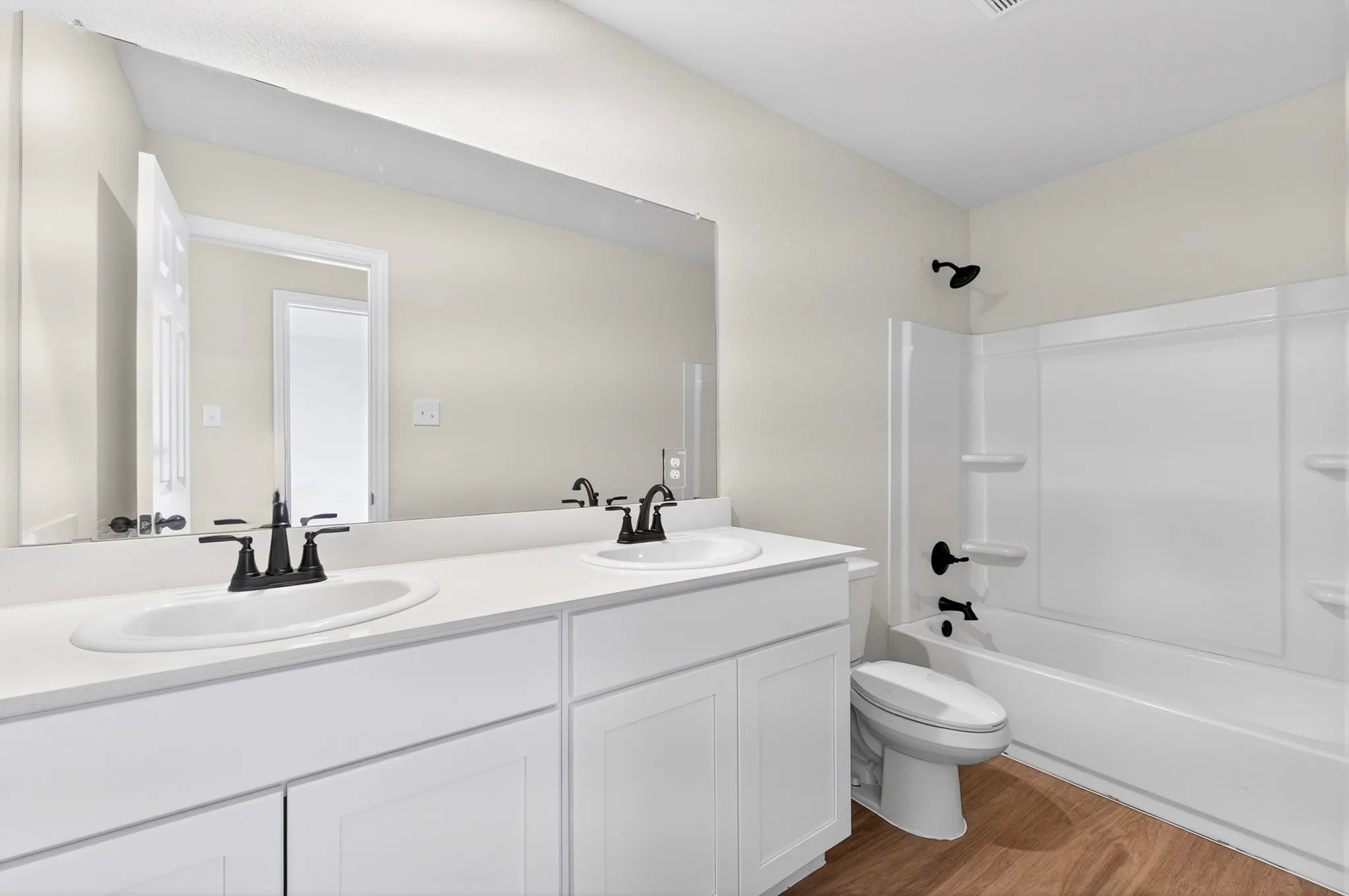 Dual vanity with white cabinetry and integrated sinks, a wide mirror, a white bathtub with a shower, and wood-finish flooring