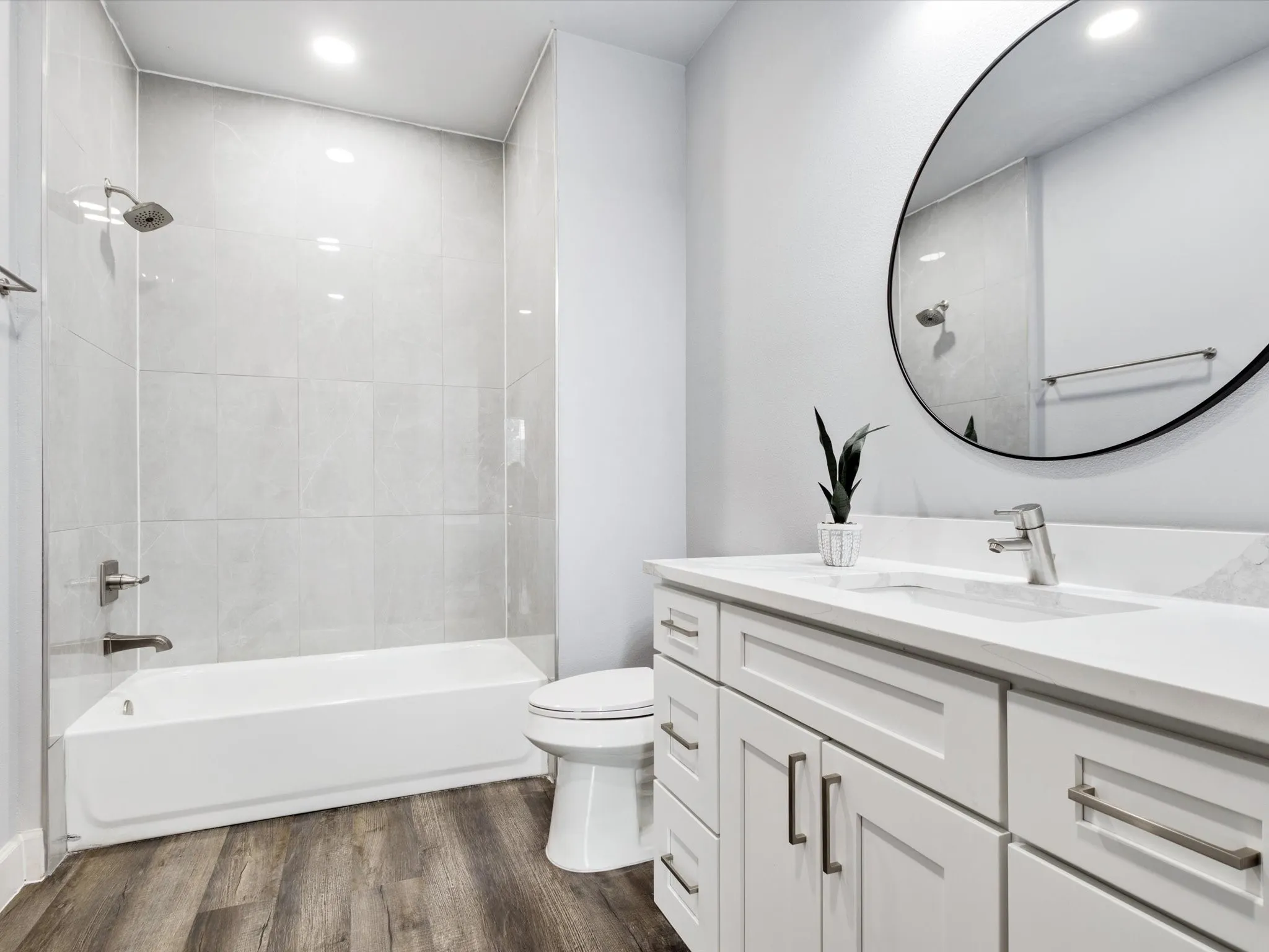 Bathroom featuring a white vanity with integrated sink, a large round mirror, wood-finish flooring, and a tiled shower with a white tub