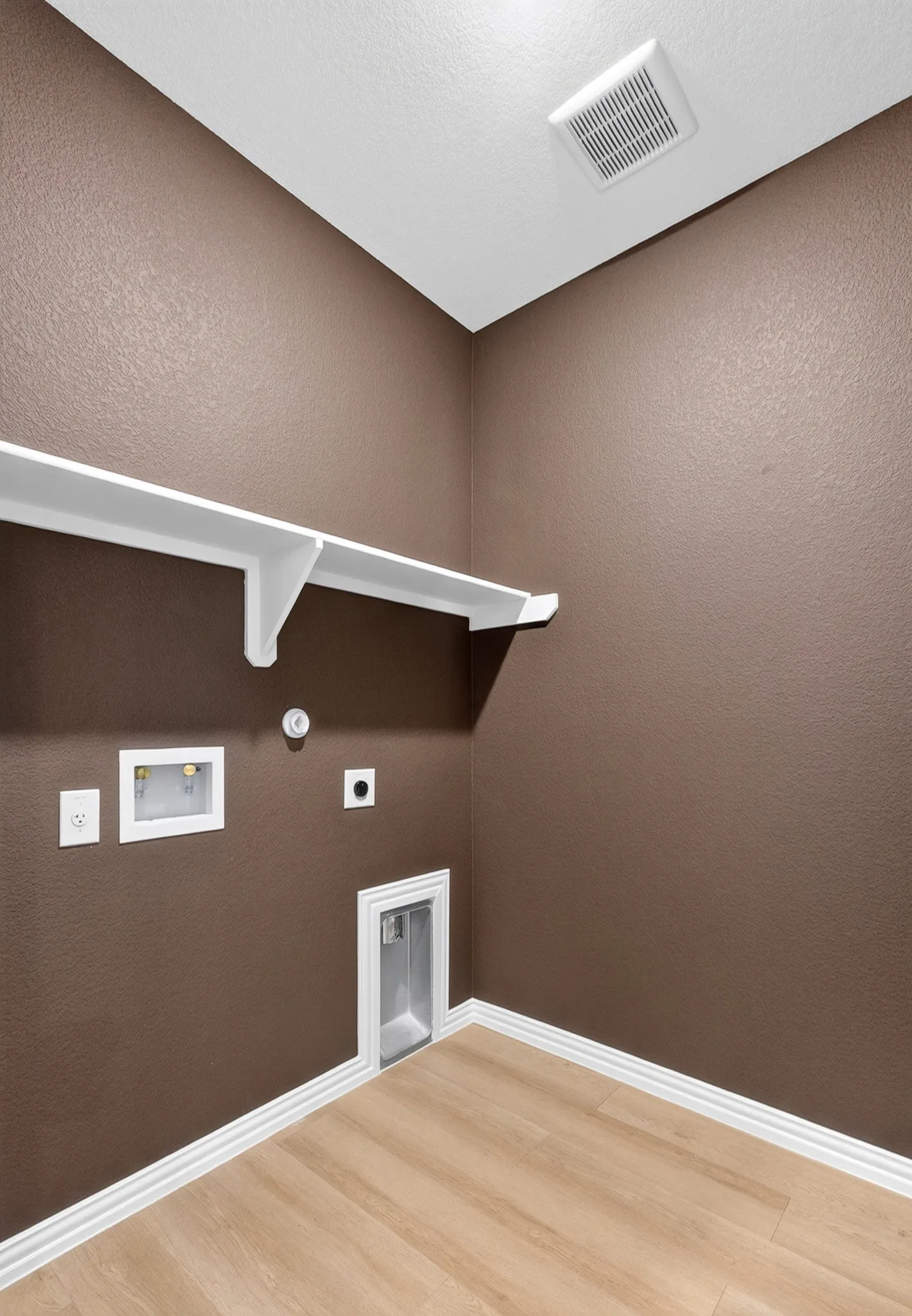 Wood-finish flooring with white baseboards, a white wall shelf, and built-in utility connections