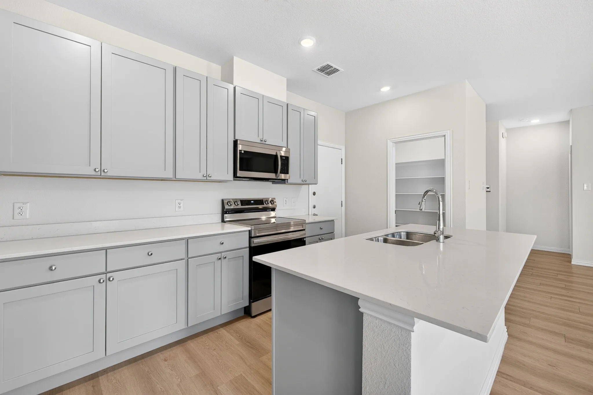 Kitchen featuring light gray cabinetry, stainless steel appliances, a center island with an integrated sink, white countertops, and wood-finish flooring