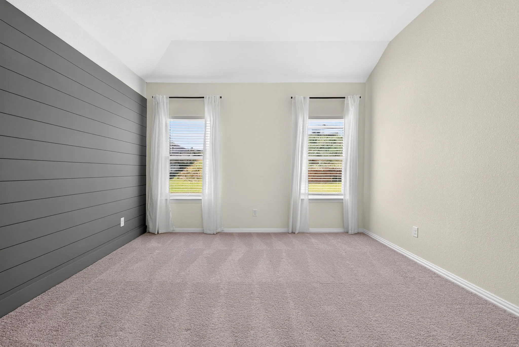 Vaulted ceiling, two windows with blinds, dark accent wall with horizontal paneling, light-toned walls, and neutral carpeting