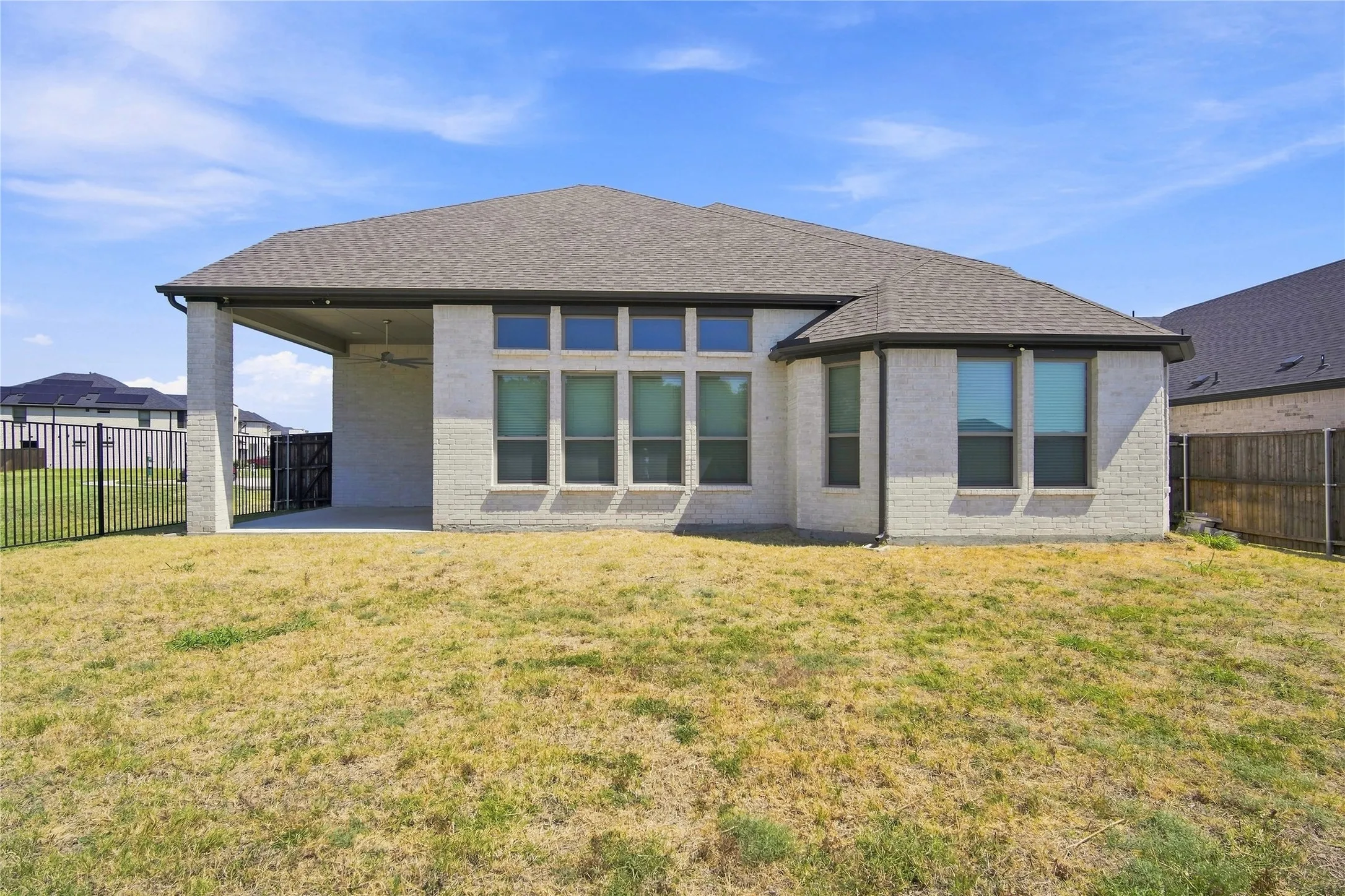 Modern residence featuring a geometric hip roof with architectural stone and stucco exterior, expansive multi-pane window array across the rear facade, manicured lawn with mature landscaping, and covered patio structure with substantial columns