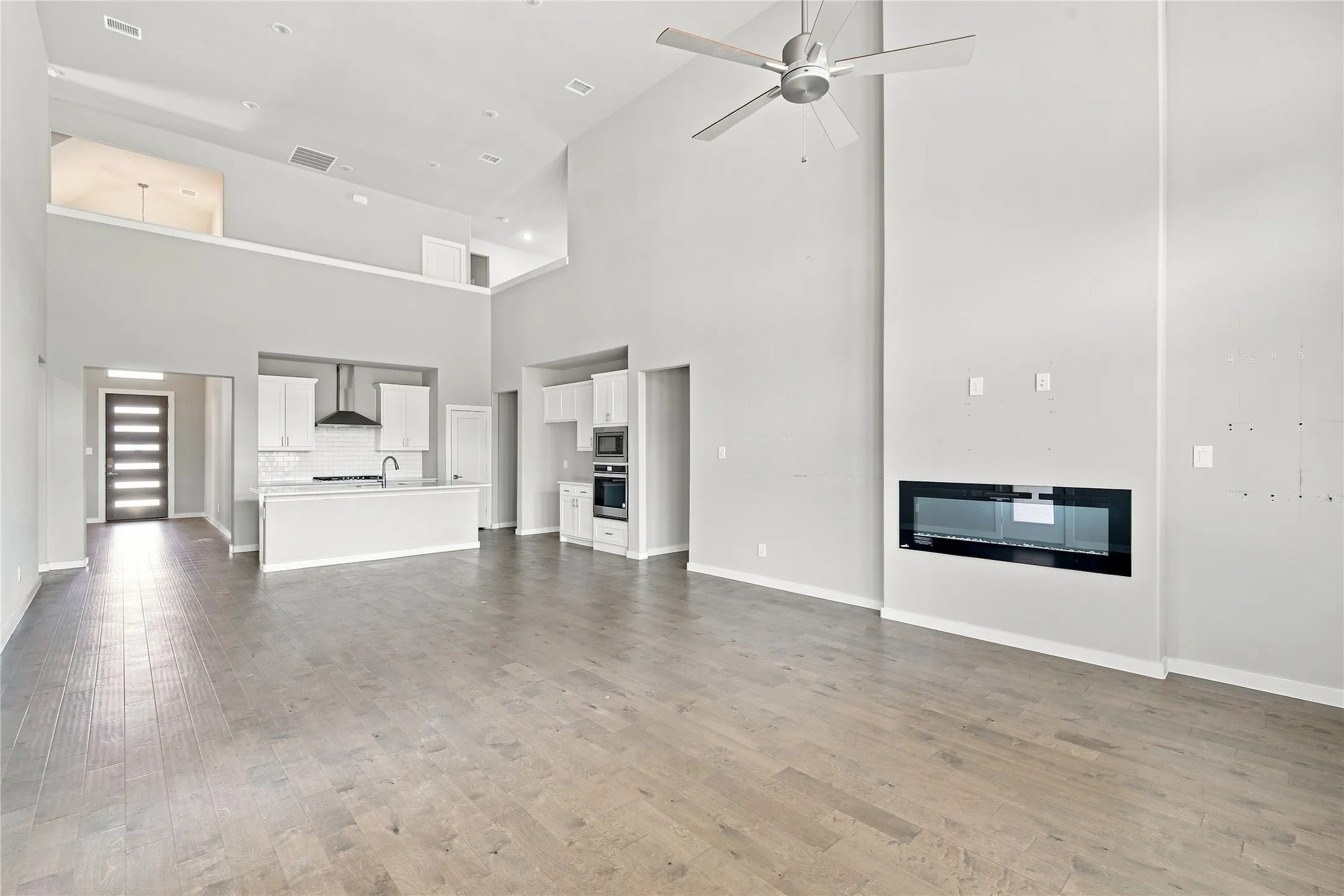 Vaulted ceilings with a ceiling fan, wood-finish flooring, built-in linear fireplace, kitchen island with sink, and white cabinetry with appliances
