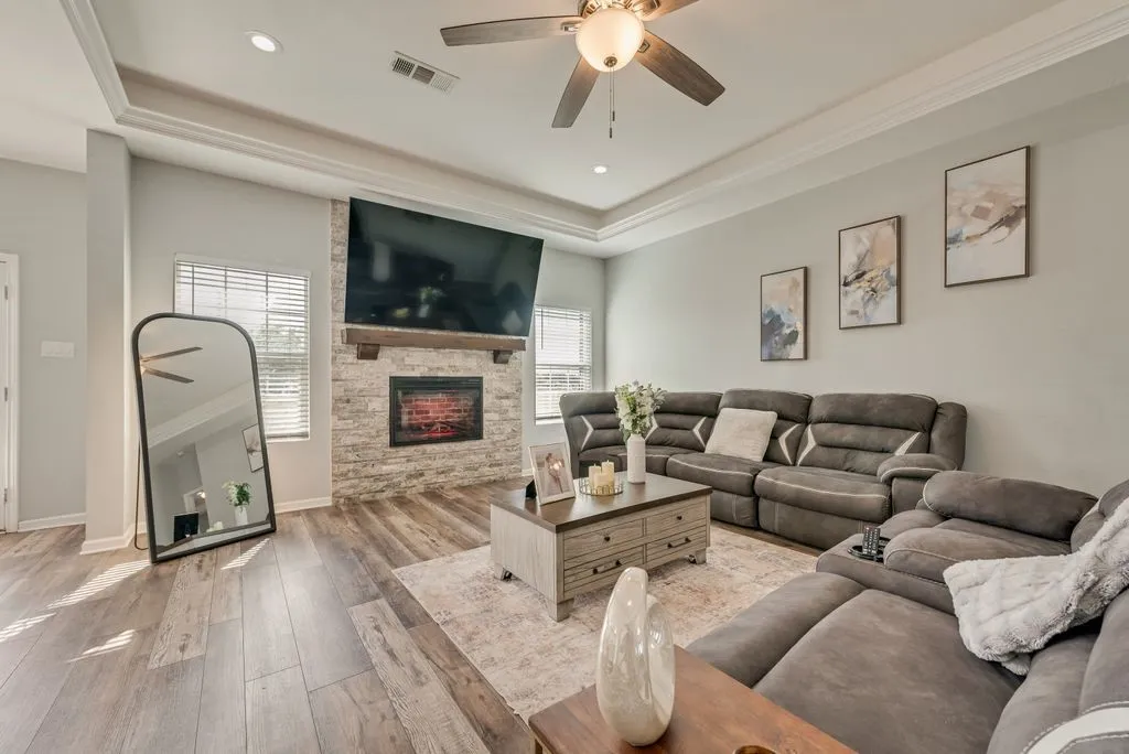 Tray ceiling with crown molding, recessed lighting, wood-finish flooring, stone-clad fireplace, and a ceiling fan with integrated lighting