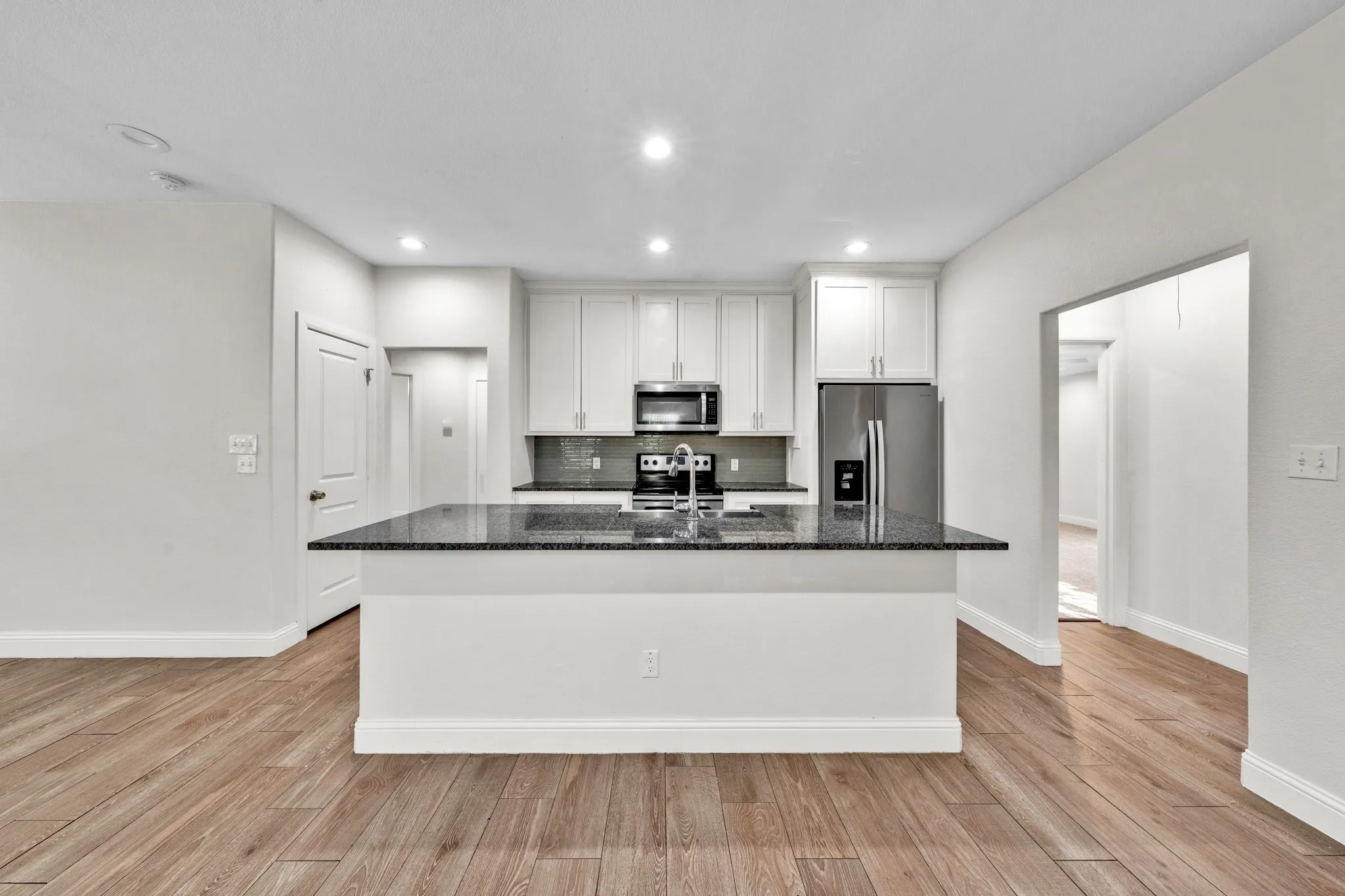 Kitchen featuring an island with a built-in sink, white cabinetry, dark speckled countertops, stainless steel appliances, and wood-finish flooring