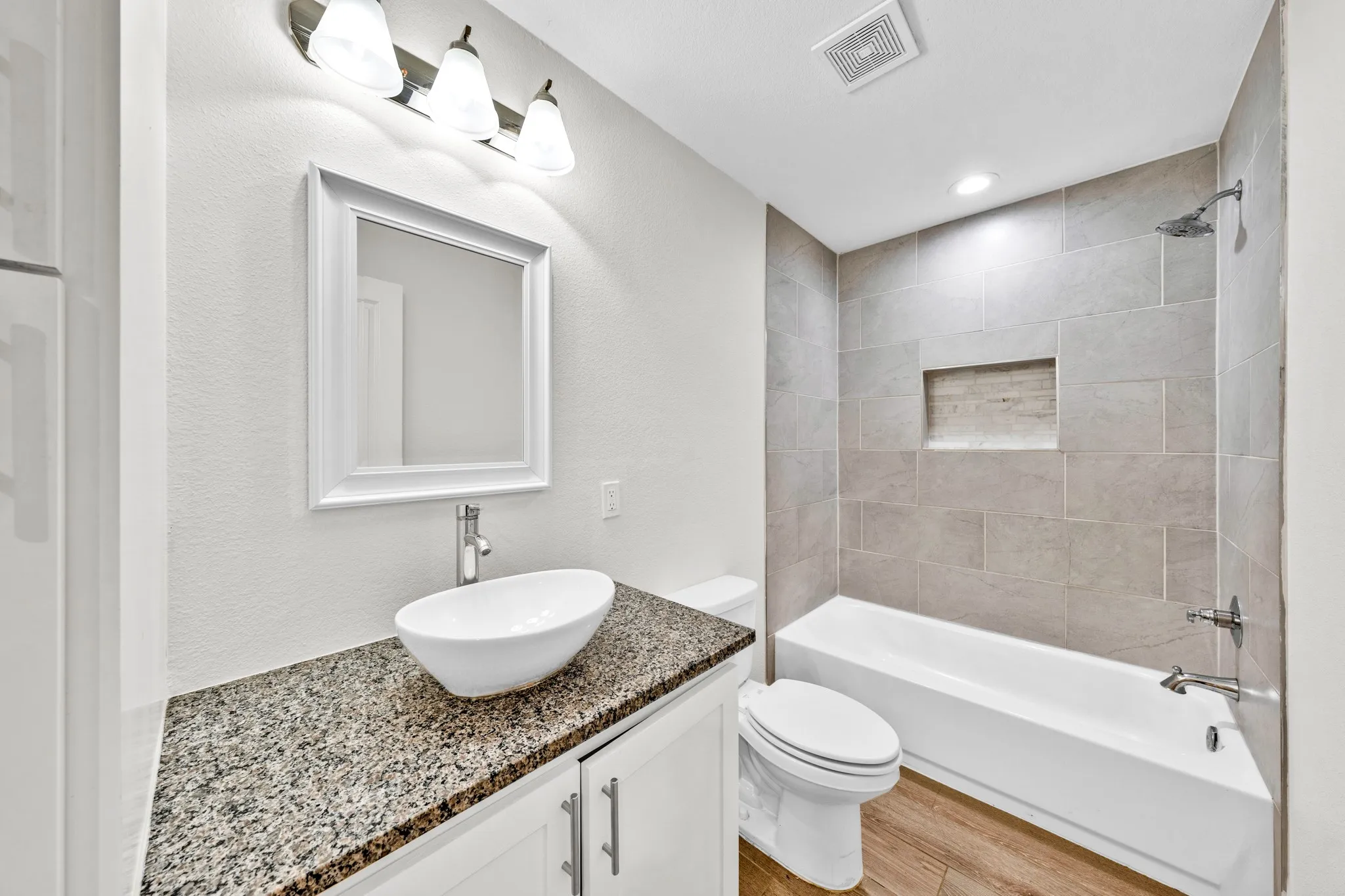 Bathroom featuring a vessel sink, granite-style countertop, white vanity cabinetry, tub with tile surround, and wood-finish flooring