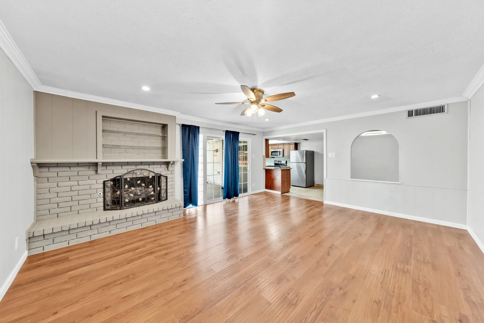 Wood-finish flooring, brick-faced fireplace with built-in shelving, ceiling fan with integrated lighting, recessed lighting, and a pass-through arched opening
