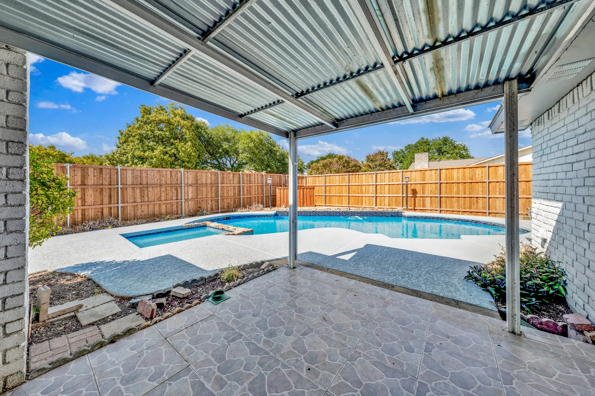 Covered patio with a tiled surface and corrugated metal roof, overlooking a backyard swimming pool