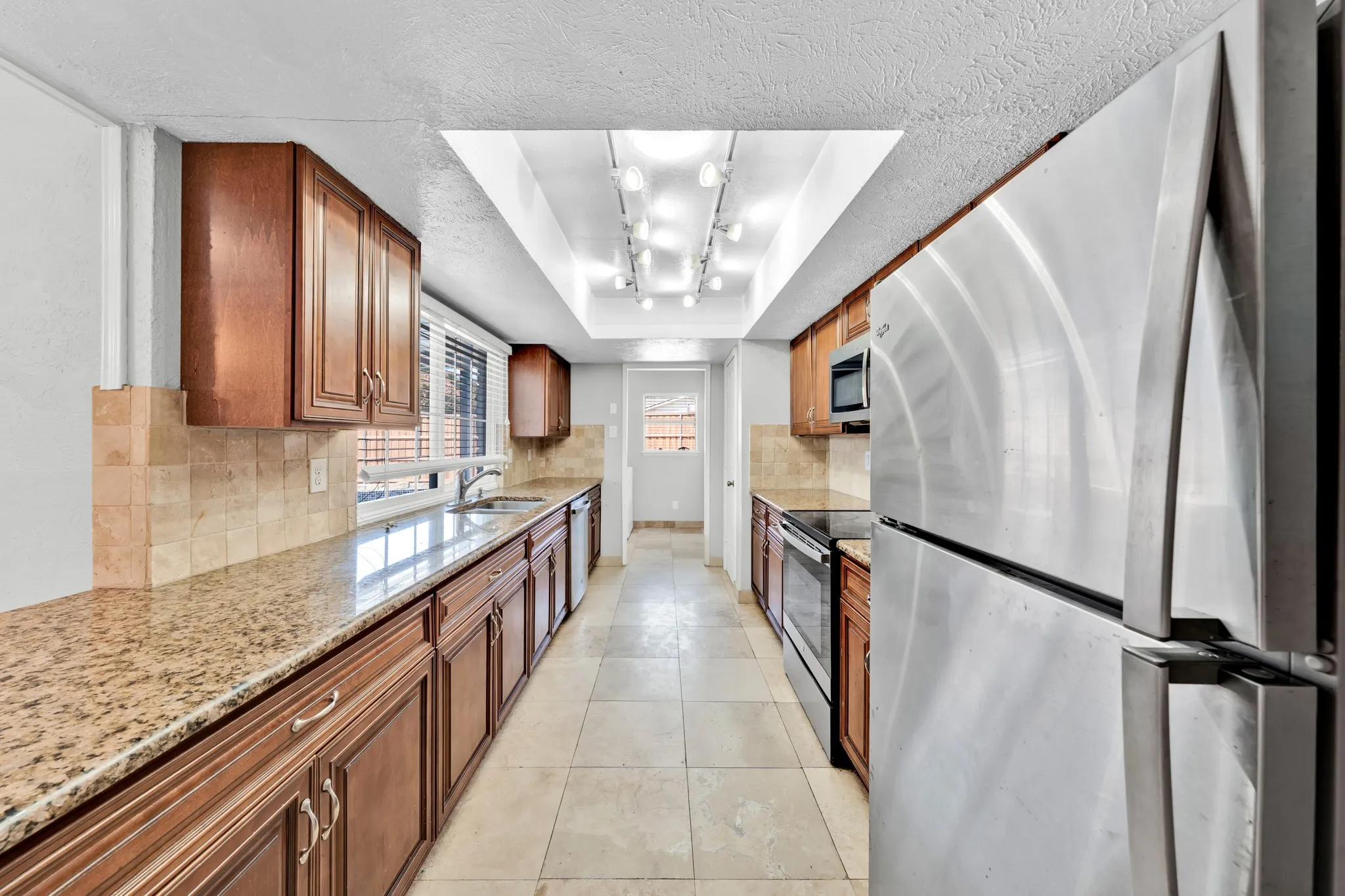 Galley kitchen featuring wood cabinetry, stone-finish countertops, tile backsplash, stainless steel appliances, and tile flooring